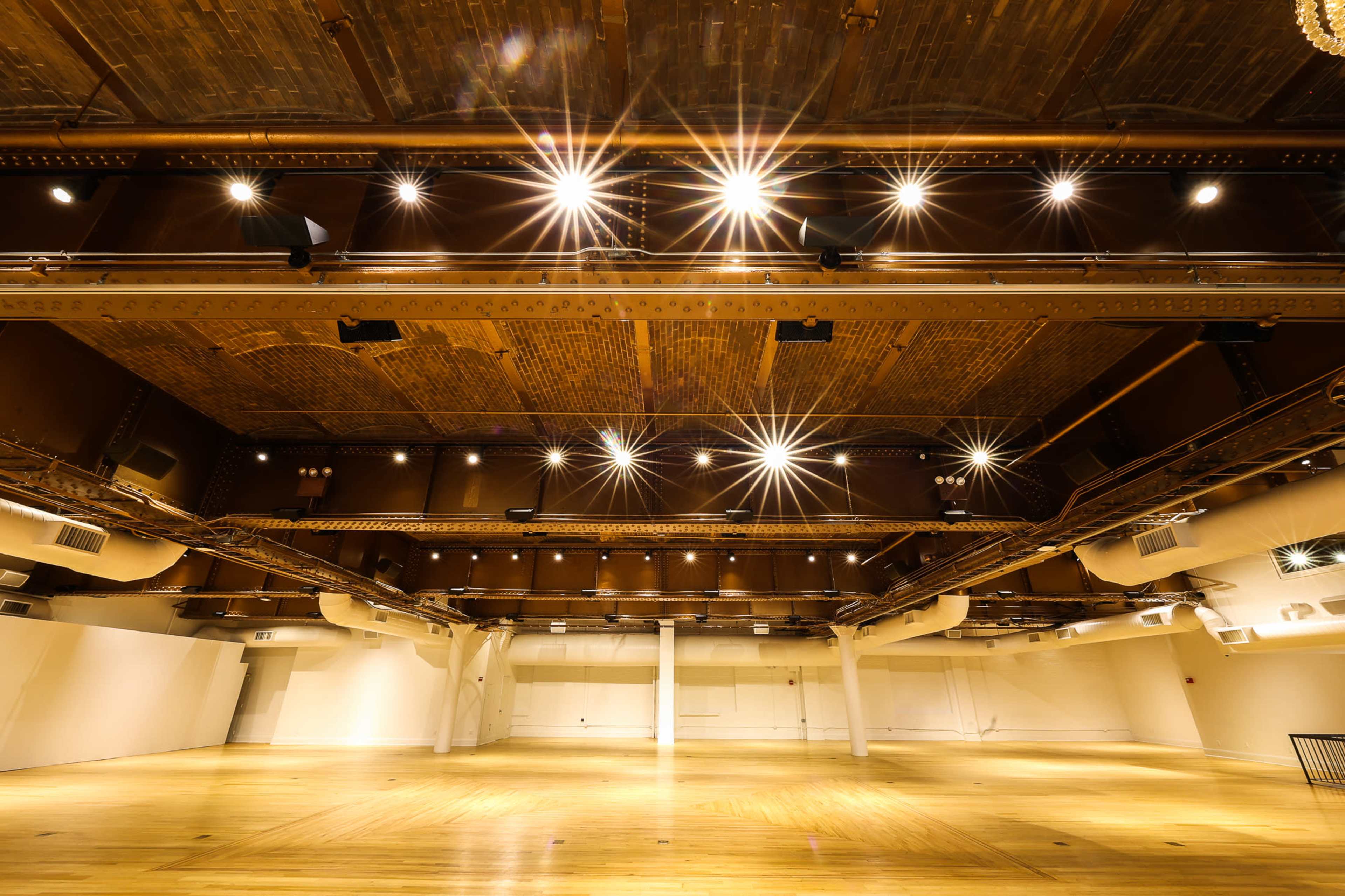 An empty hall with wooden floors and a high ceiling illuminated by bright overhead lights.