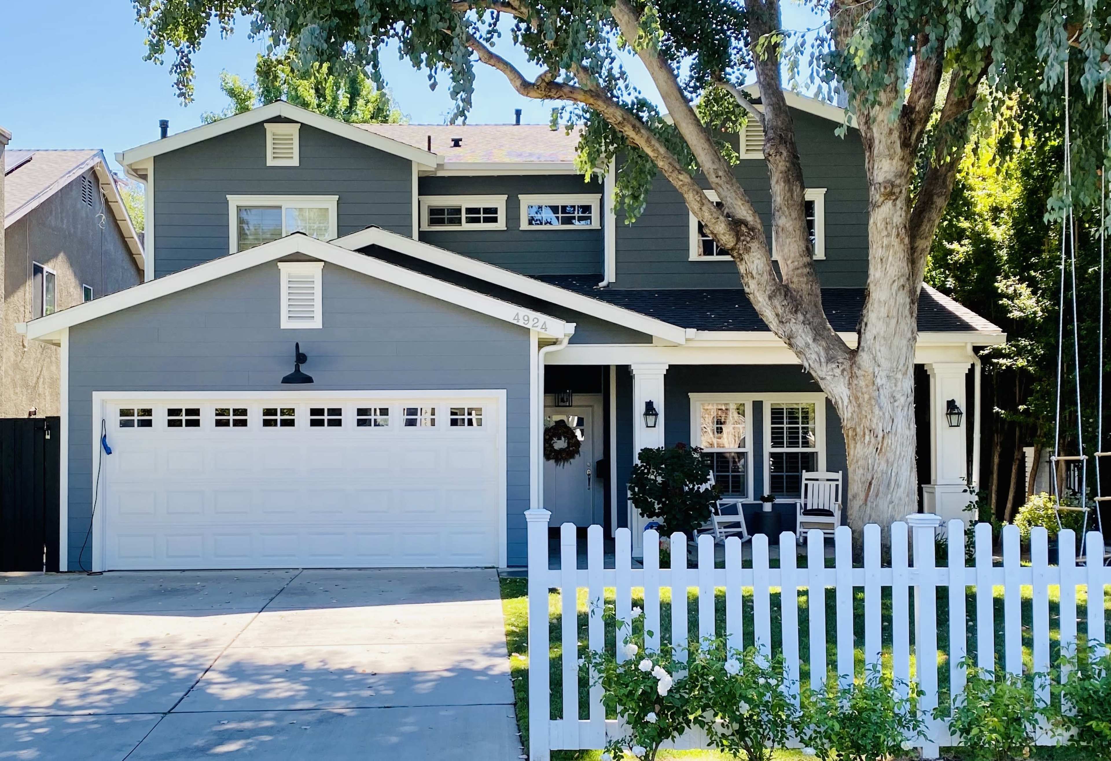 A two-story gray house with a white picket fence, a concrete driveway, and a large tree in the front yard.