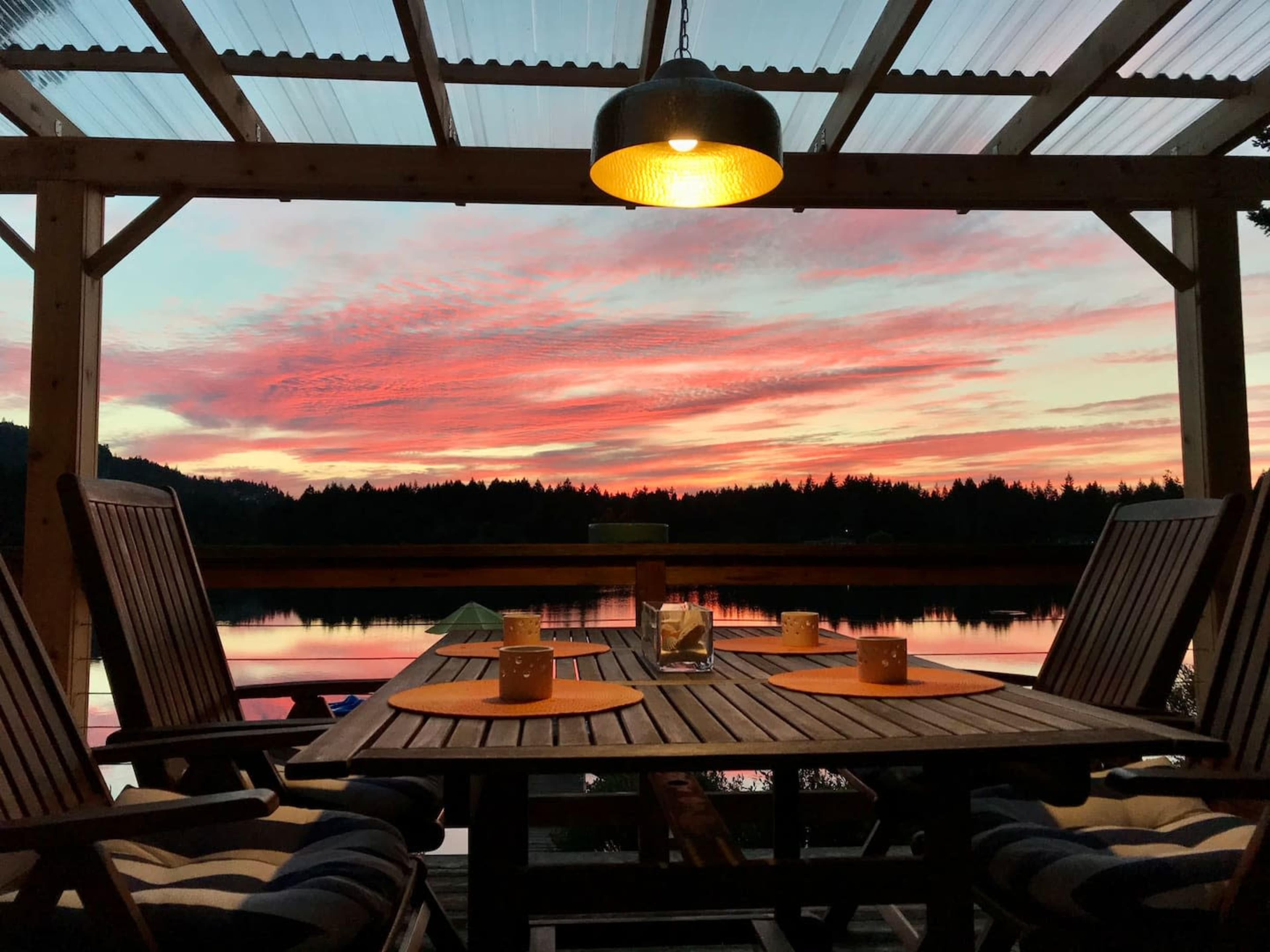 A wooden table and chairs are set under a covered patio, overlooking a calm lake at sunset, with vibrant colors in the sky reflected on the water.