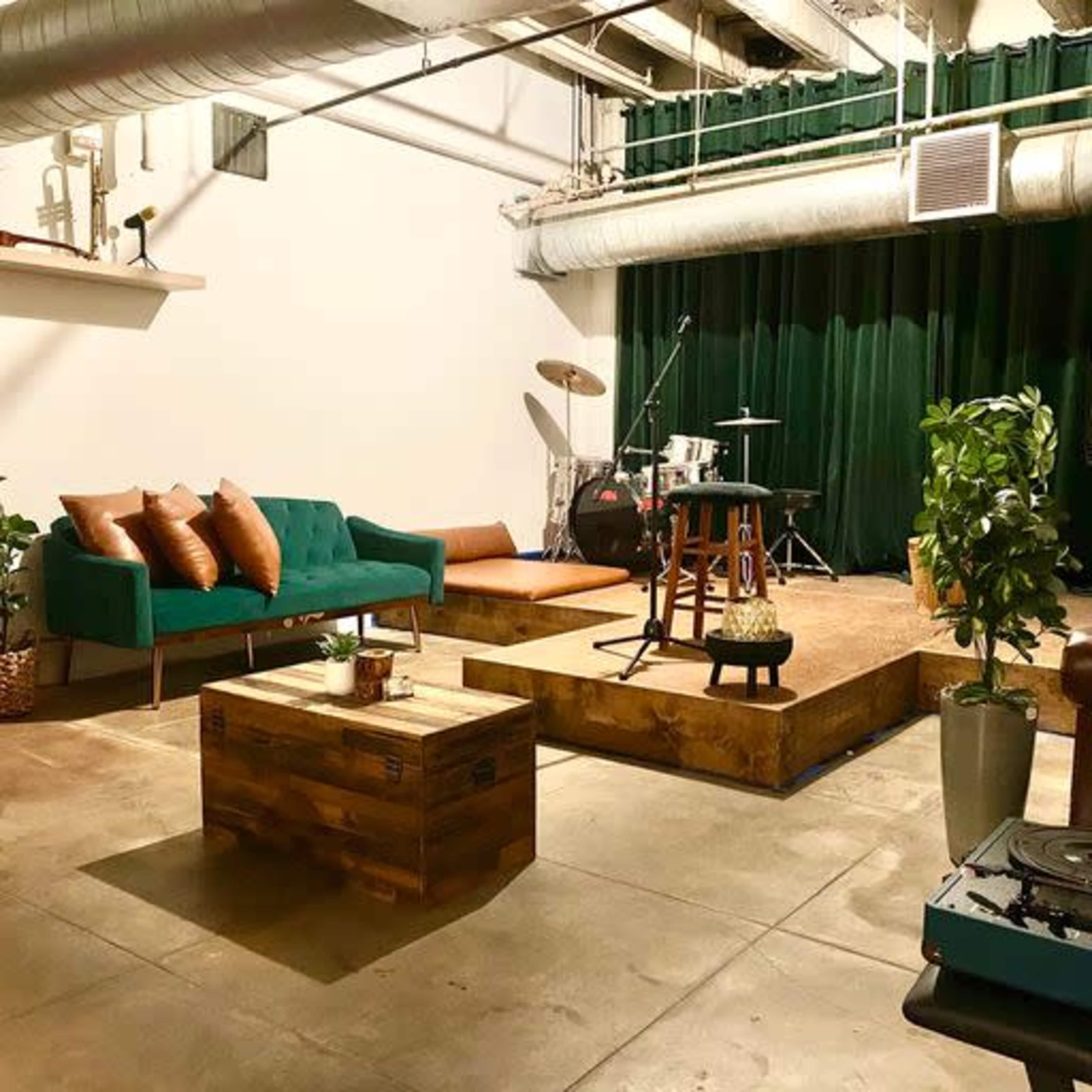A cozy lounge area with a green couch and a wooden coffee table, a small stage with musical instruments, and a backdrop of green curtains.