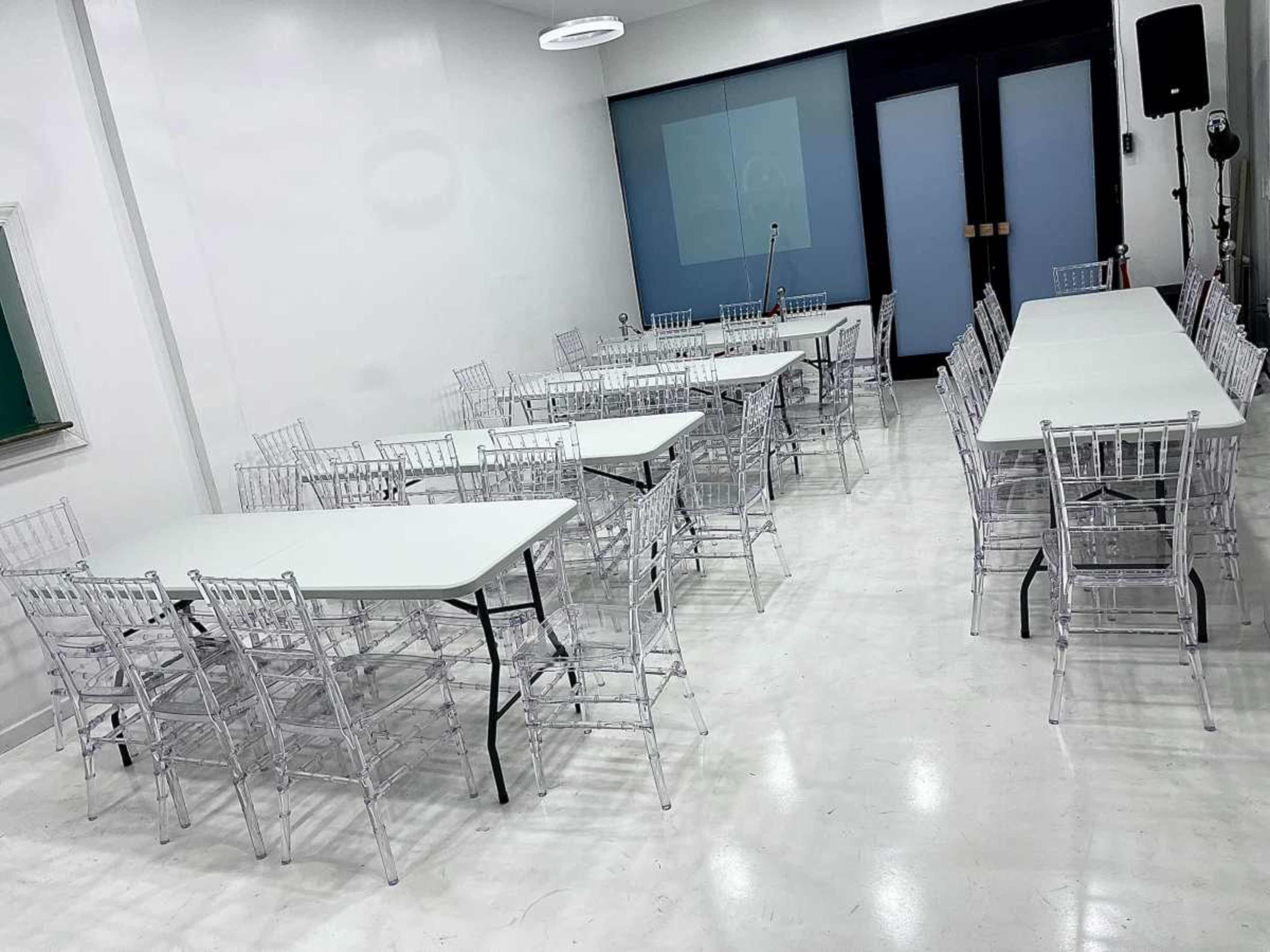 The image shows a spacious meeting room arranged with multiple clear plastic tables and chairs, set up in a rectangular configuration.
