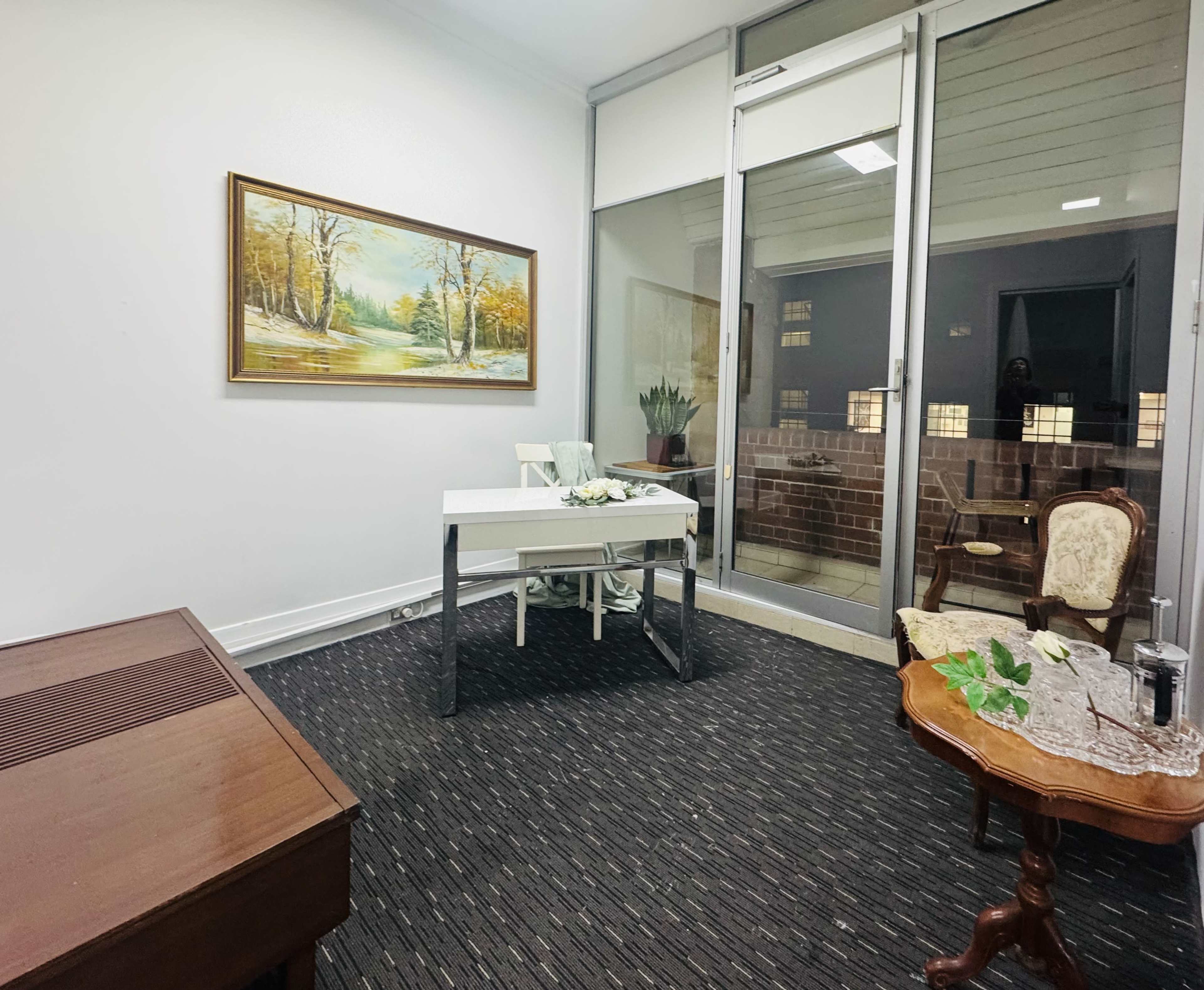 The image shows a small, well-lit office space featuring a table with a floral arrangement, a wooden desk, and a vintage armchair, with a landscape painting on the wall.