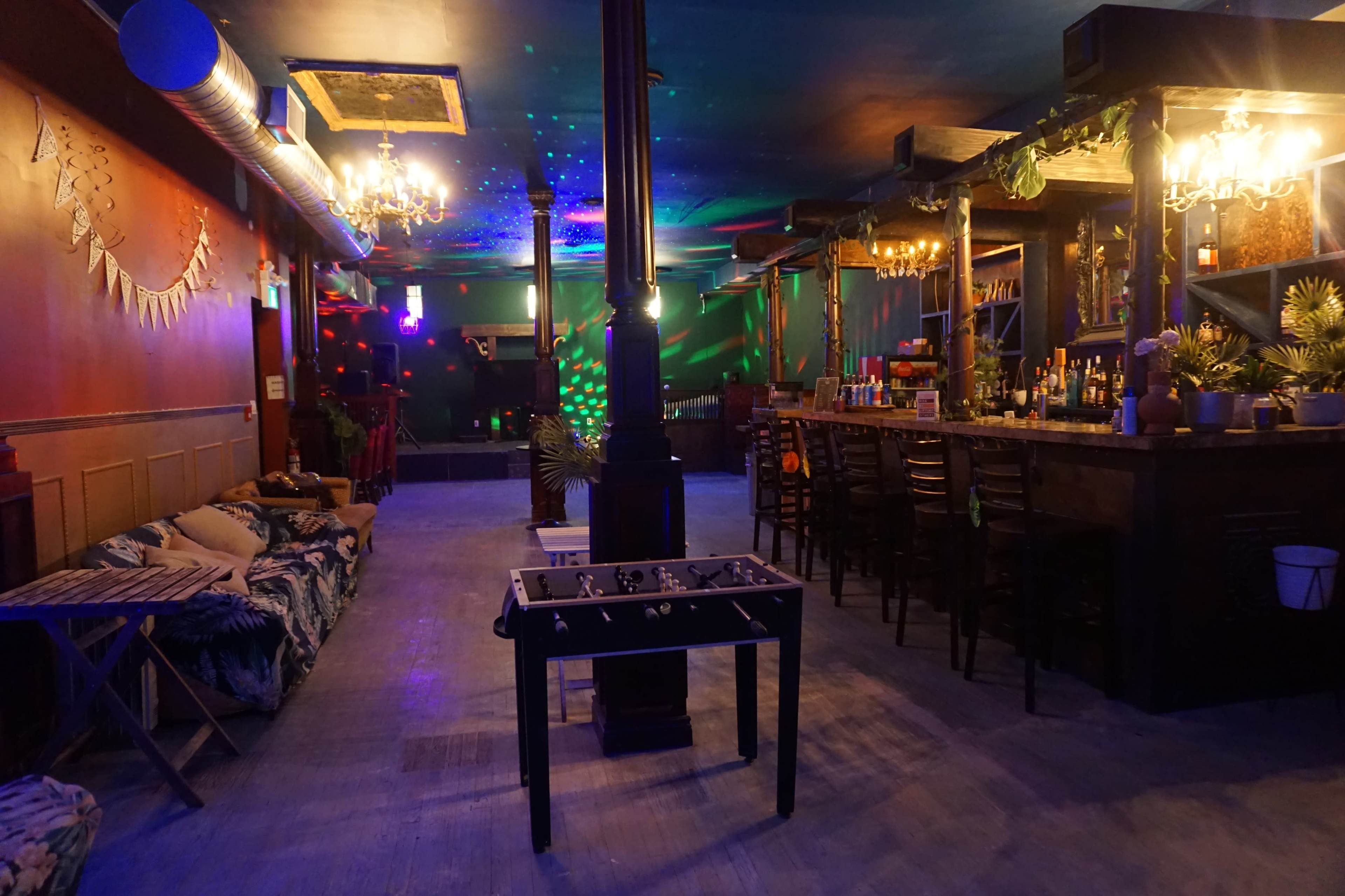 An indoor venue with a bar area, seating arrangements, a foosball table, and colorful lights illuminating the space.