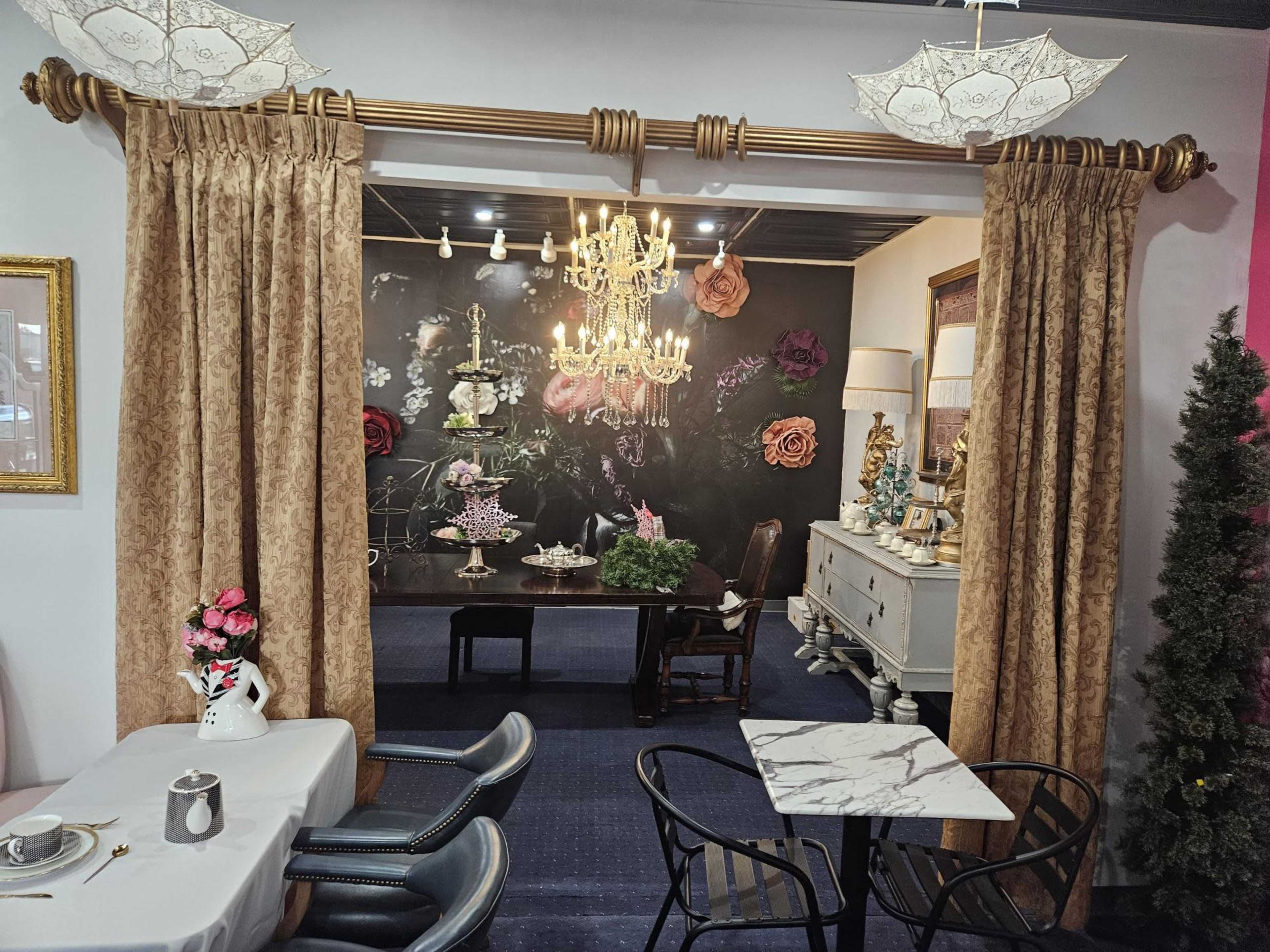 A dining area features elegant curtains, a chandelier, and floral wall art, leading into an adjoining space with decorative furnishings.