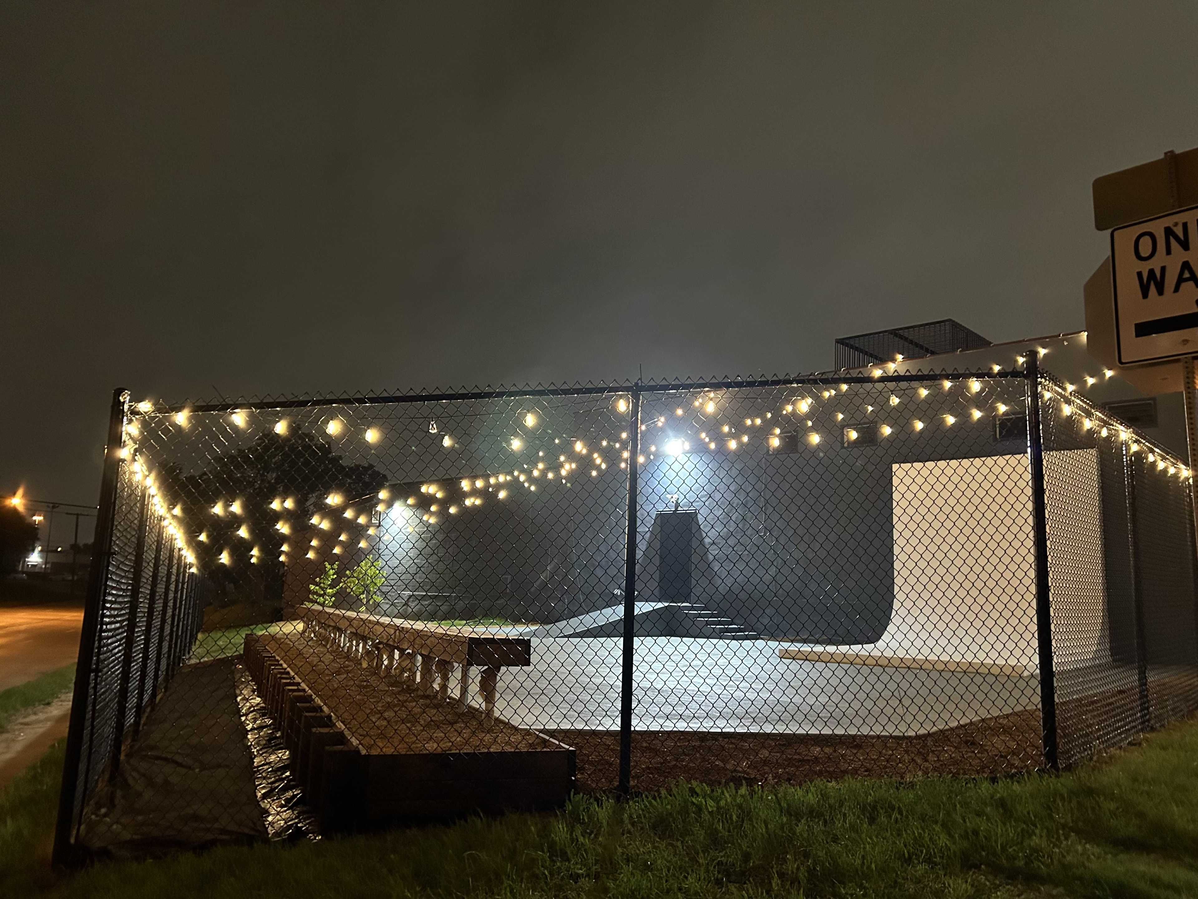 A fenced skate park at night is illuminated by string lights, with a ramp and wooden seating area.
