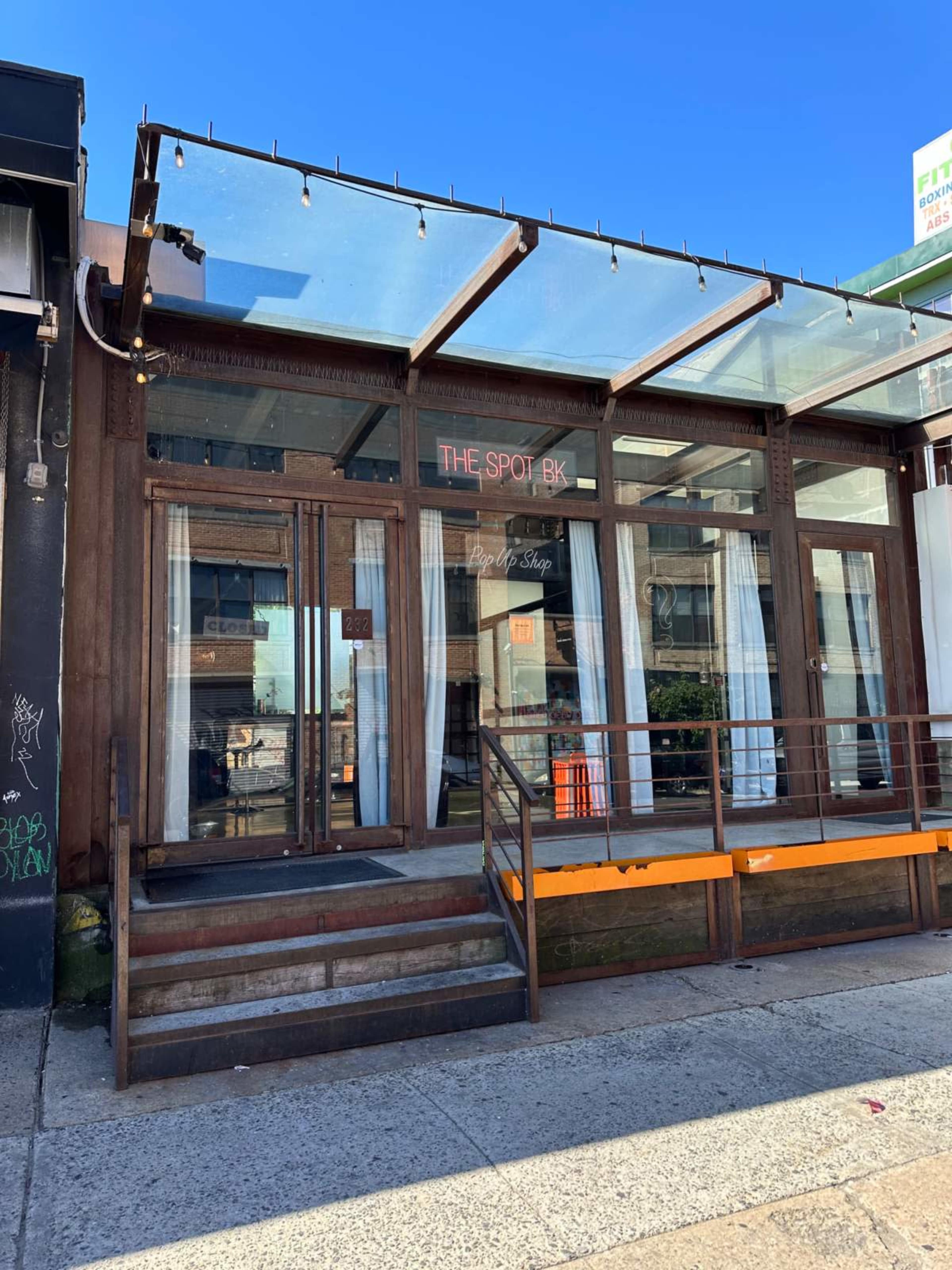 The image shows the entrance of a restaurant called "The Spot BK," with large windows, a wooden deck, and a clear awning.