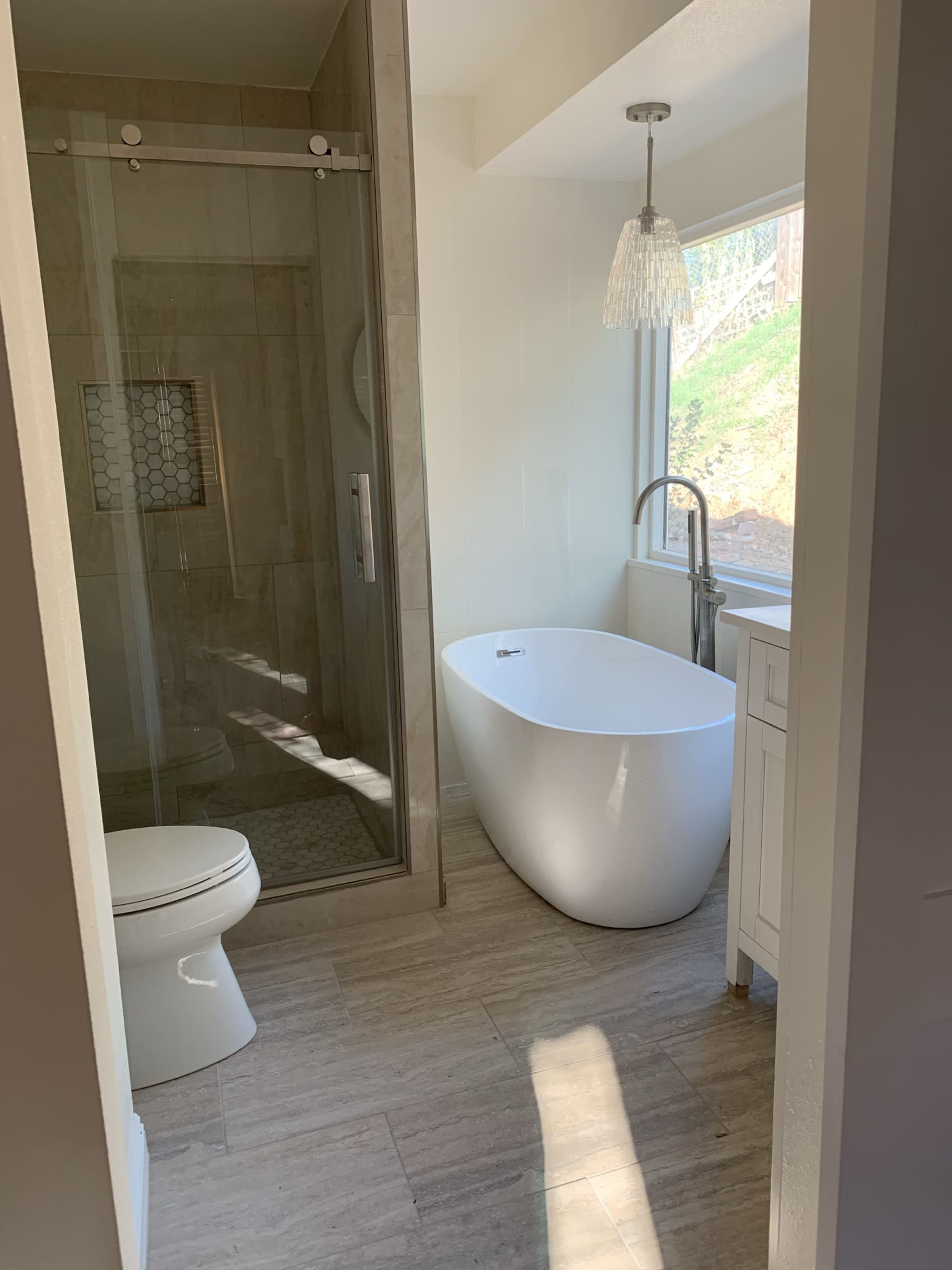 The image shows a modern bathroom featuring a glass shower enclosure, a freestanding white bathtub, and a window with natural light.