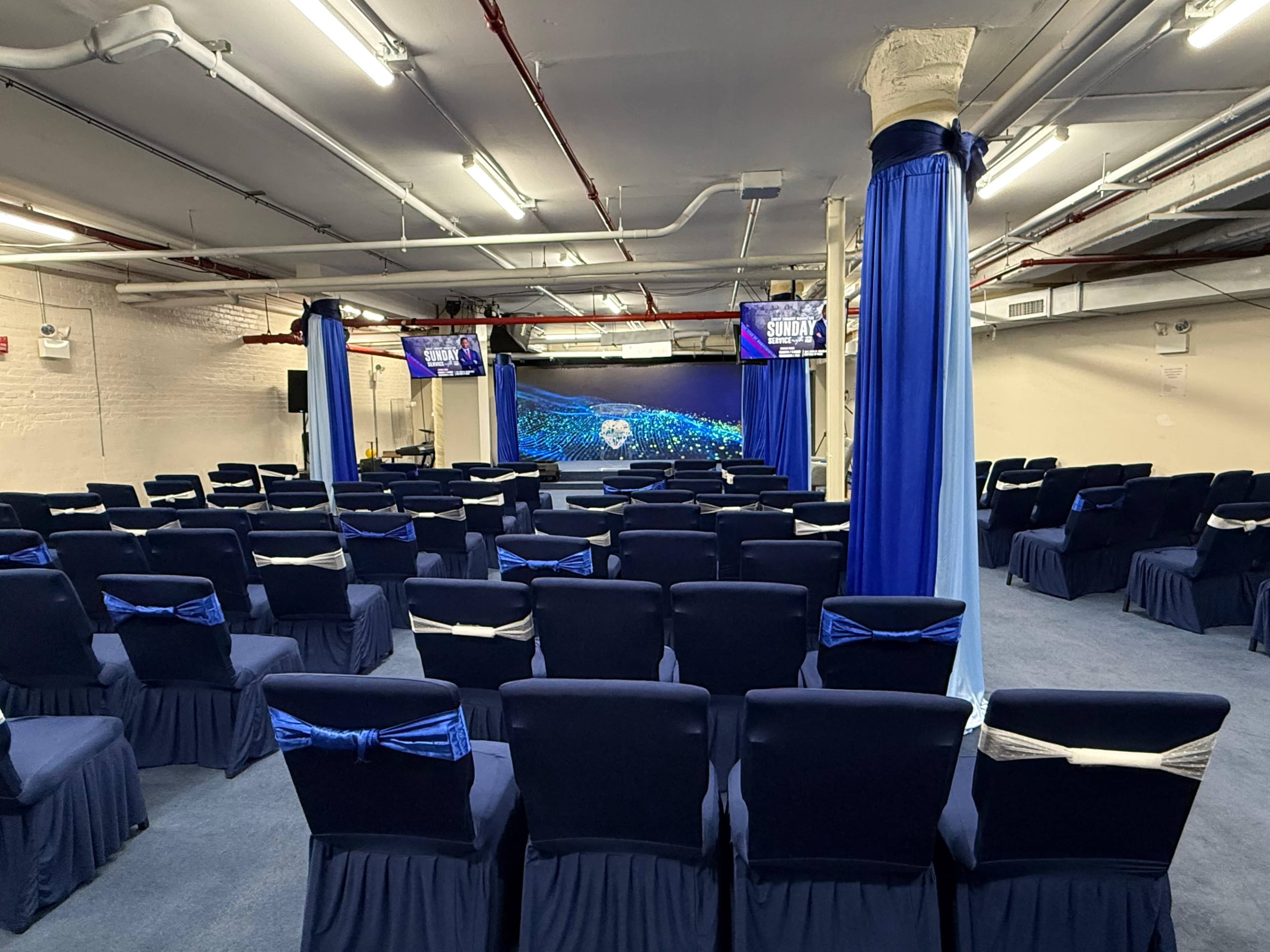The image shows an empty conference room arranged with rows of chairs covered in blue fabric, with a stage at the front displaying a digital backdrop.