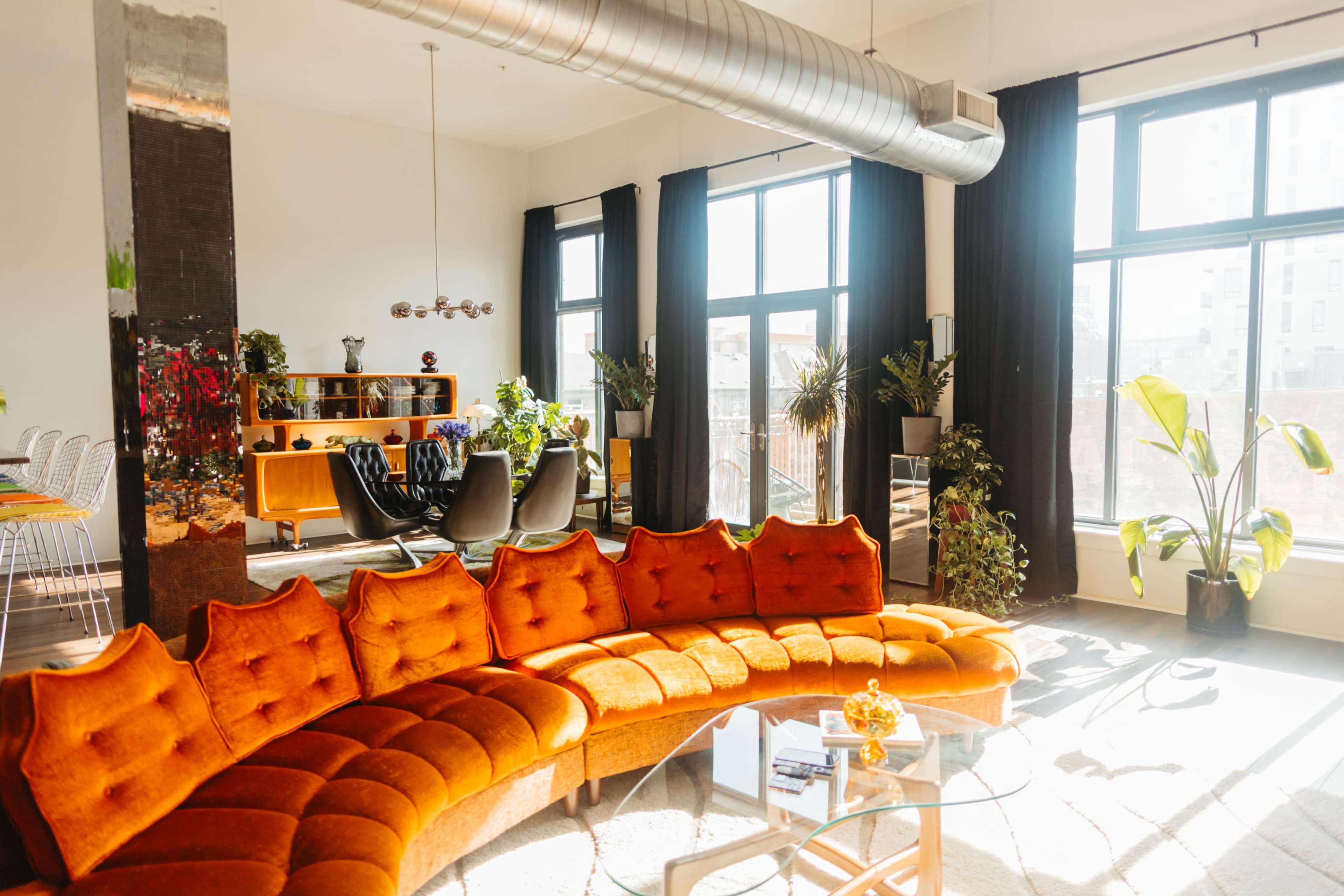 A modern living space featuring a curved orange sofa, large windows allowing natural light, and various plants and furniture arranged throughout the room.