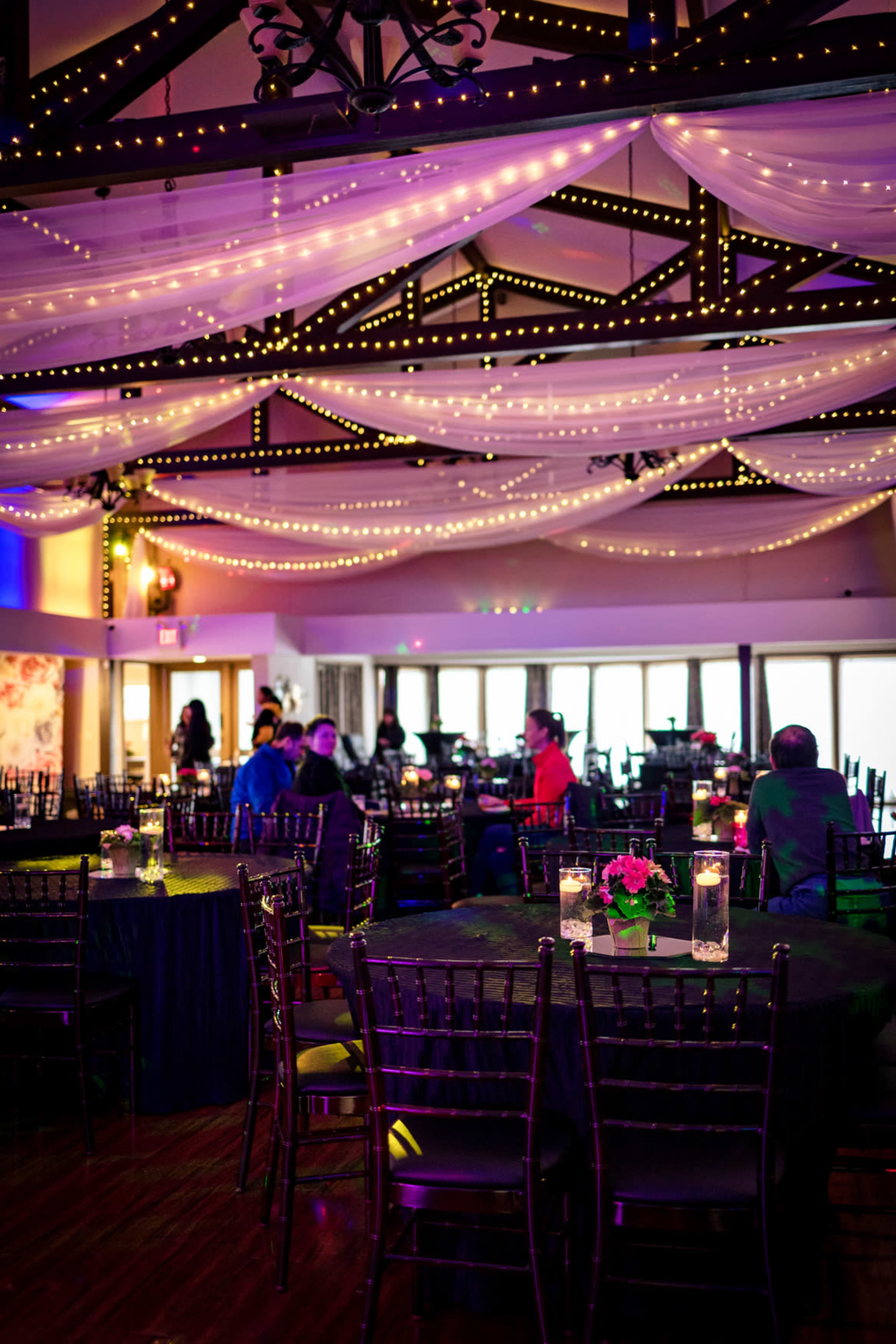 The interior space features round tables with black tablecloths, surrounded by chairs, adorned with soft lighting and draped fabric overhead.