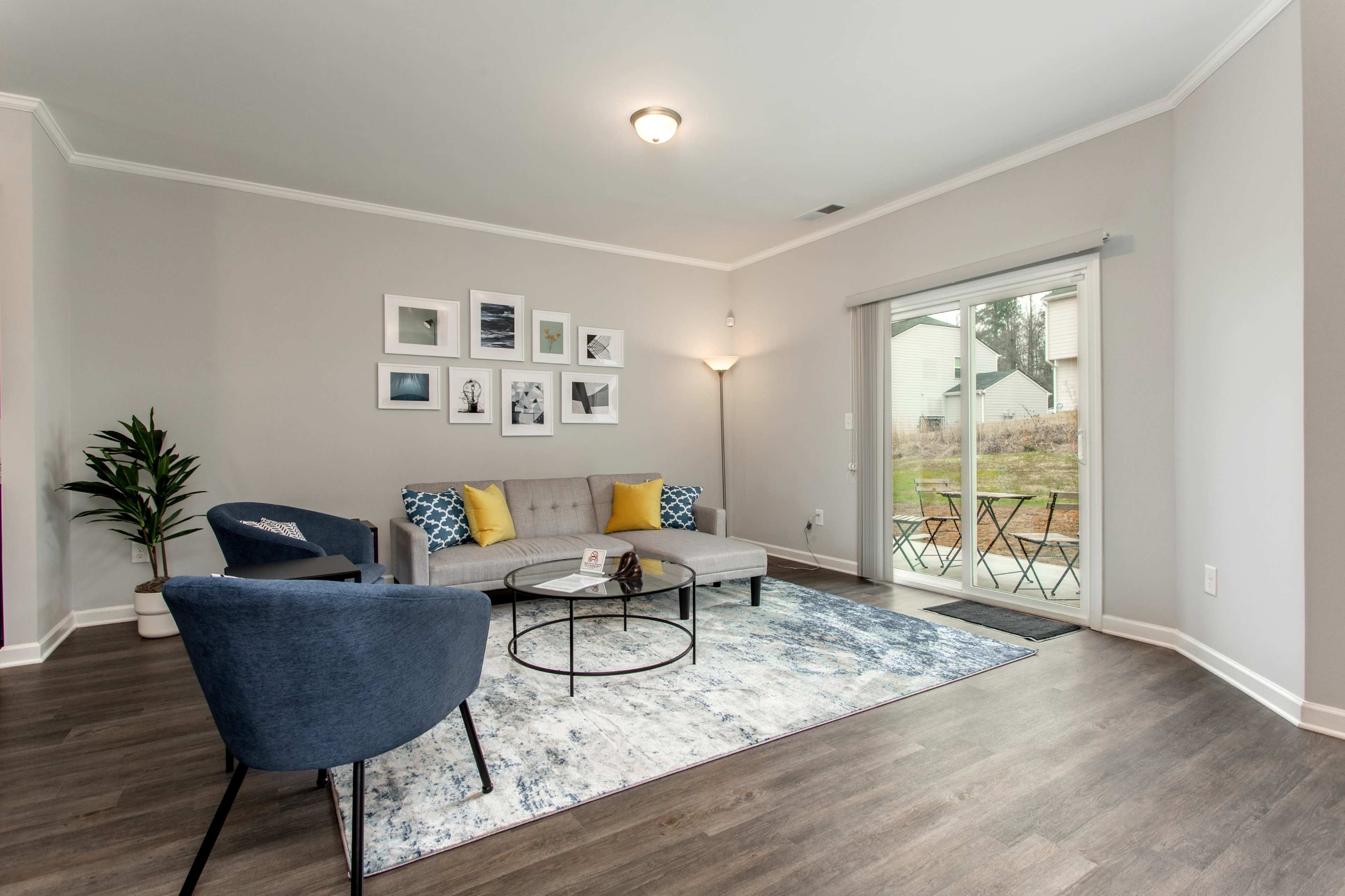 A living room features a gray sofa, two blue chairs, a coffee table, and a large area rug, with a wall of framed pictures and sliding glass doors leading to an outdoor space.