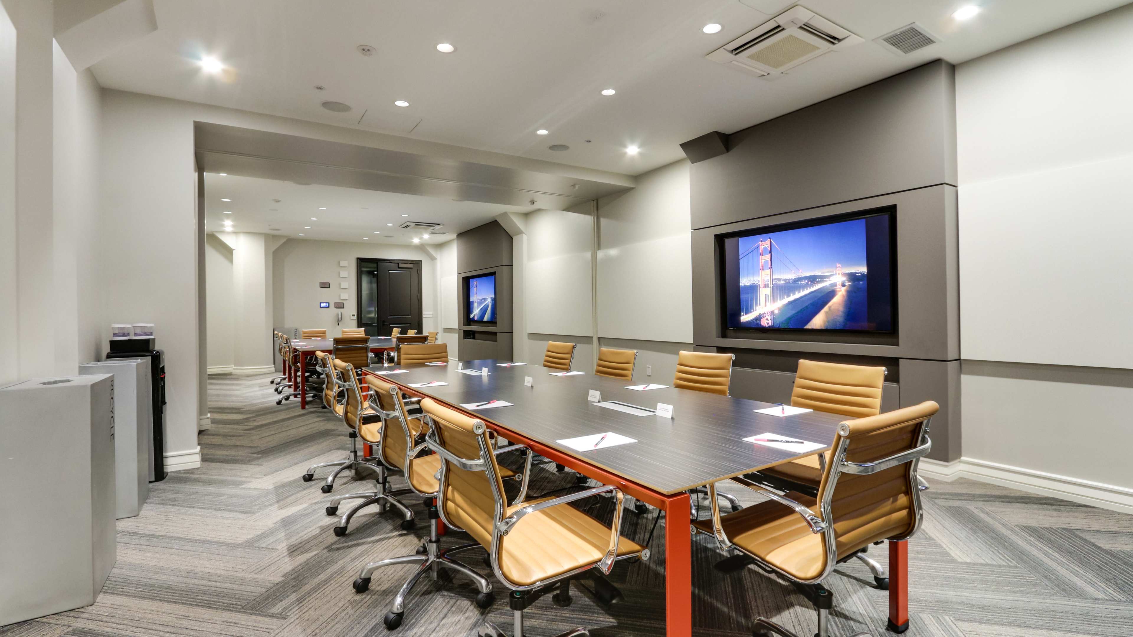 The image shows a modern conference room featuring a long table with several chairs, two screens displaying visuals, and contemporary decor.