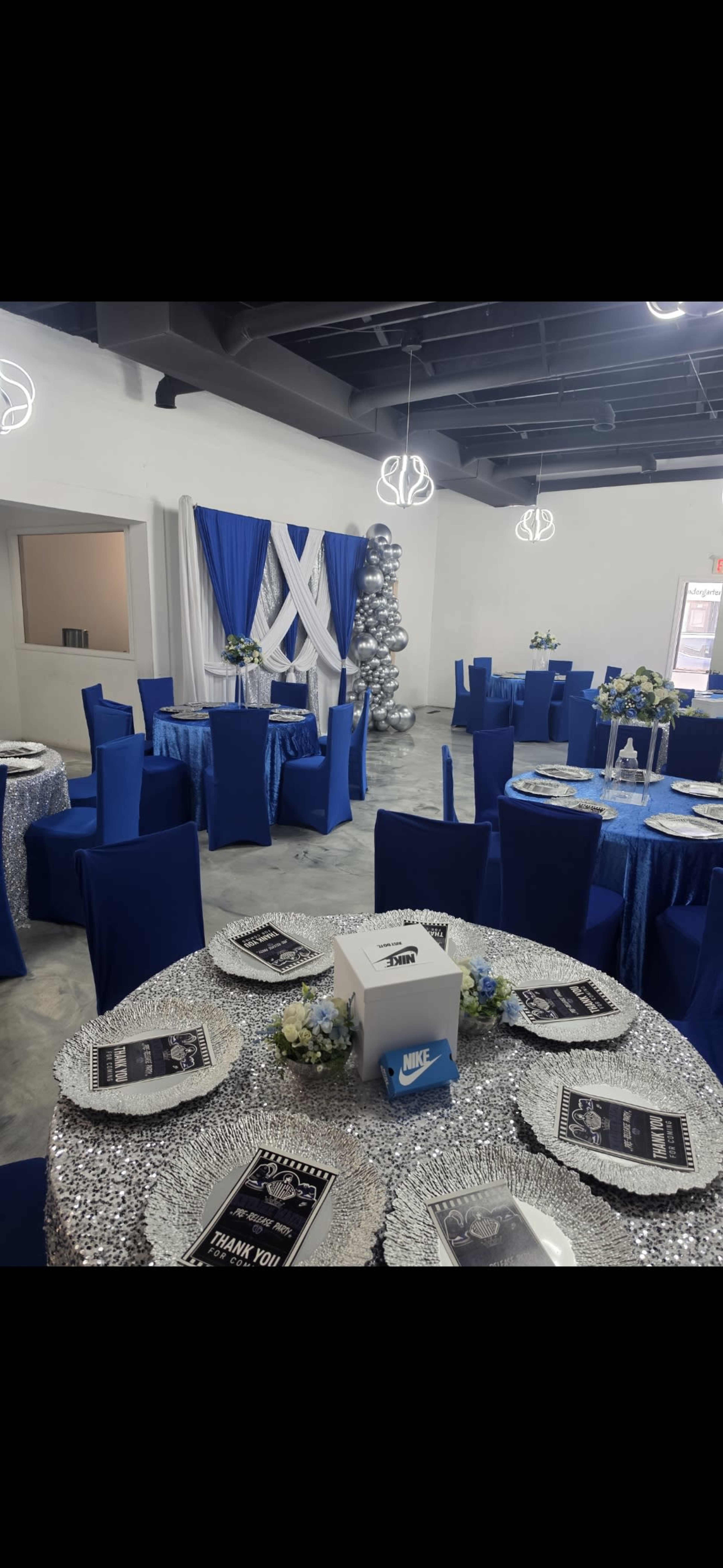 The image shows a decorated event space with blue and silver table settings, featuring sparkling tablecloths and floral centerpieces.