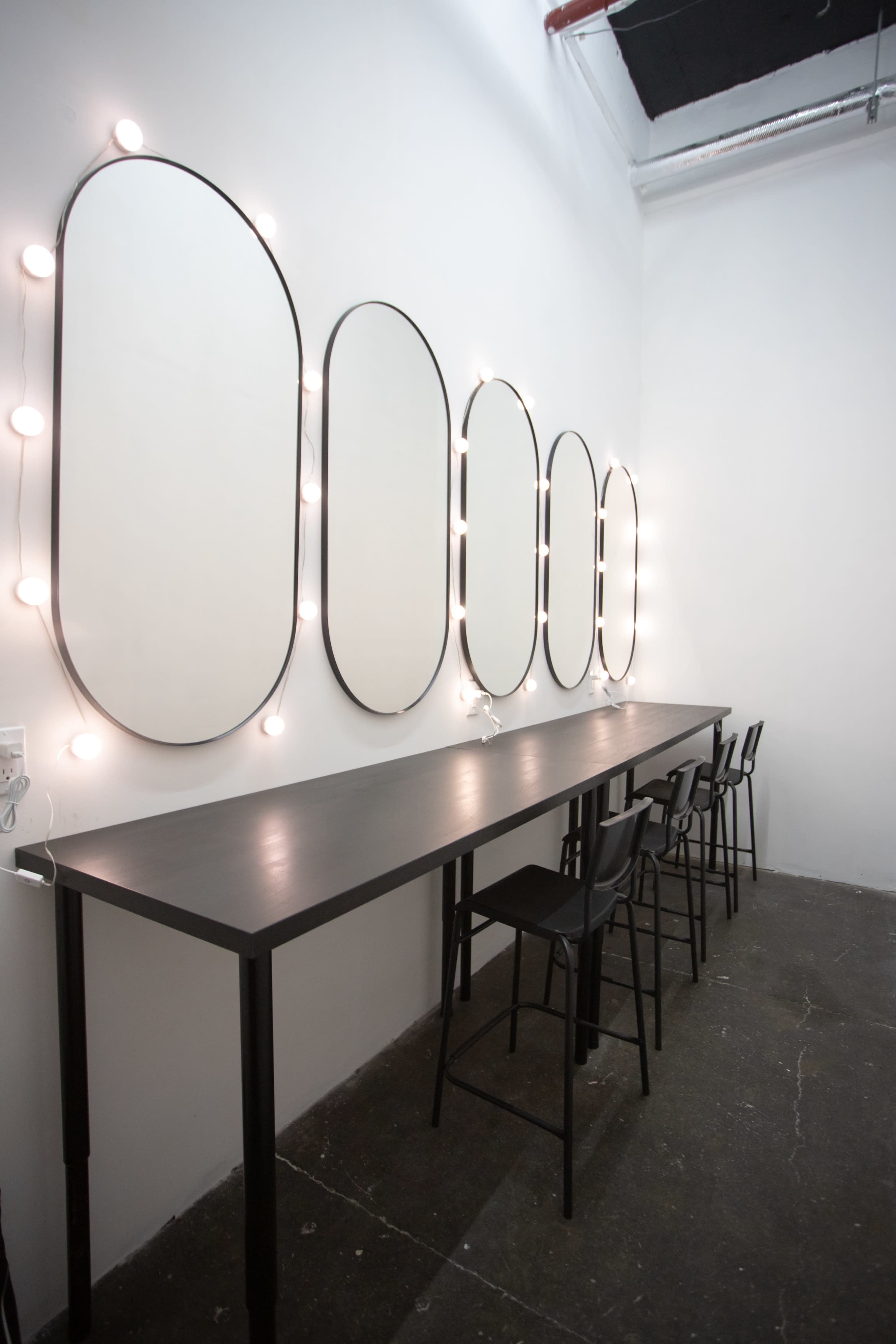 A minimalist grooming area with five oval mirrors framed in black and illuminated by round bulbs above a long black table and several black stools.