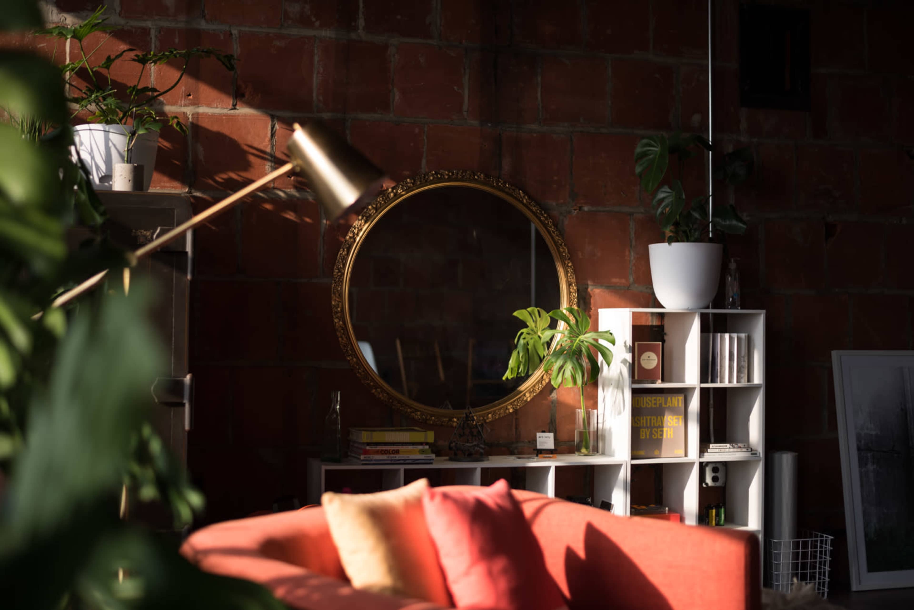 A cozy living room features a gold-framed mirror, potted plants, and a bright orange couch, all illuminated by natural light.
