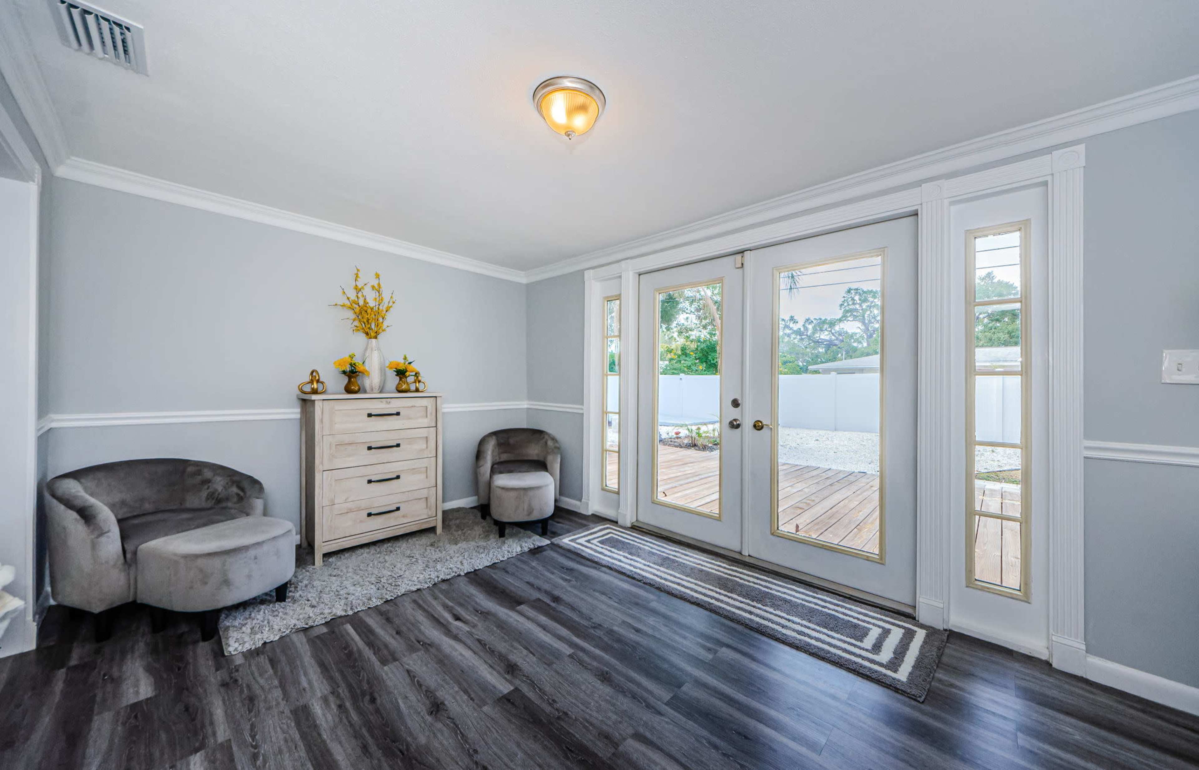 A modern entryway with a neutral-colored wall, a wooden dresser, two gray chairs, and double doors leading to an outdoor deck.