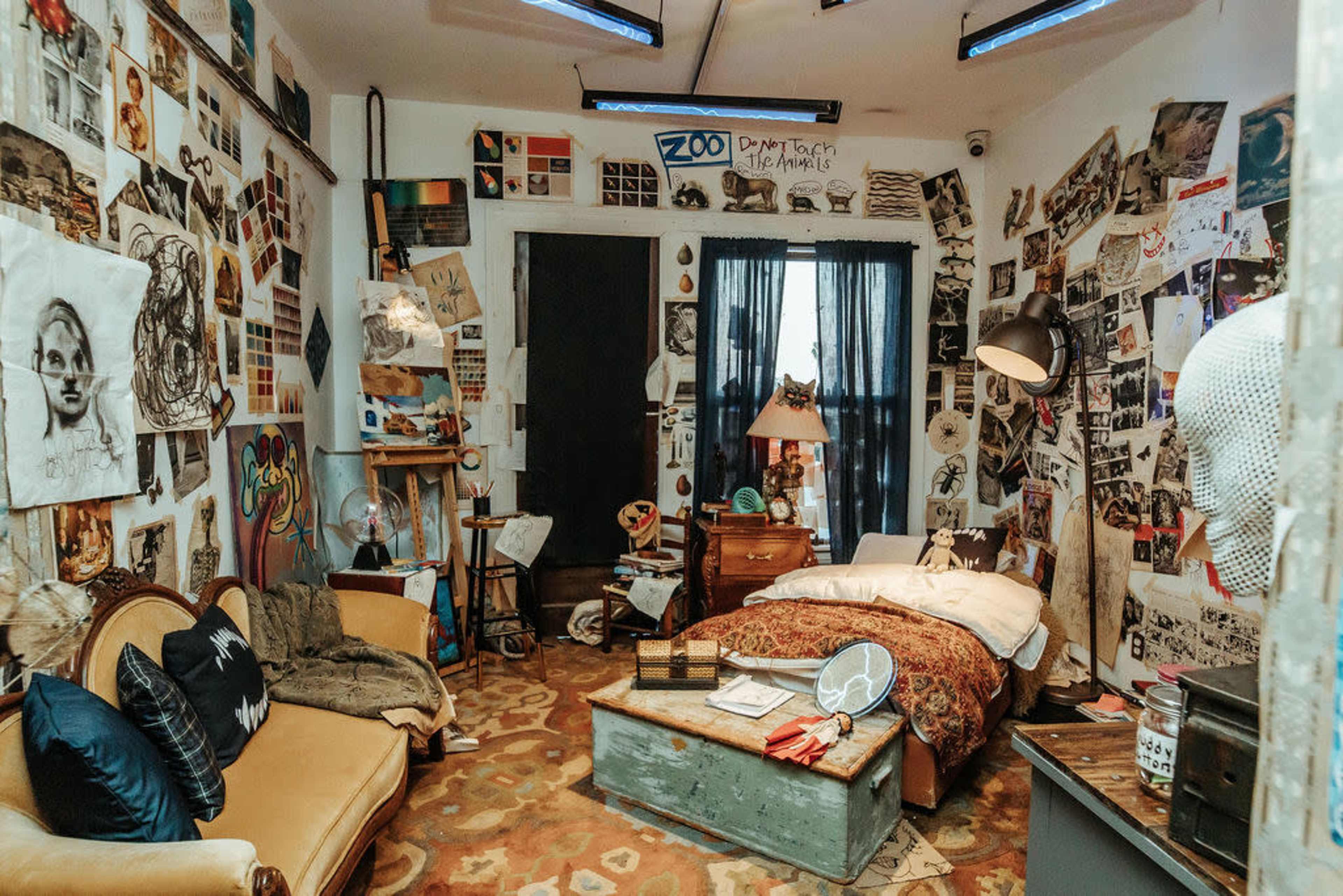 The image shows a cluttered bedroom filled with various artworks, sketches, and photographs adorning the walls, and featuring a bed, a sofa, and scattered furniture.
