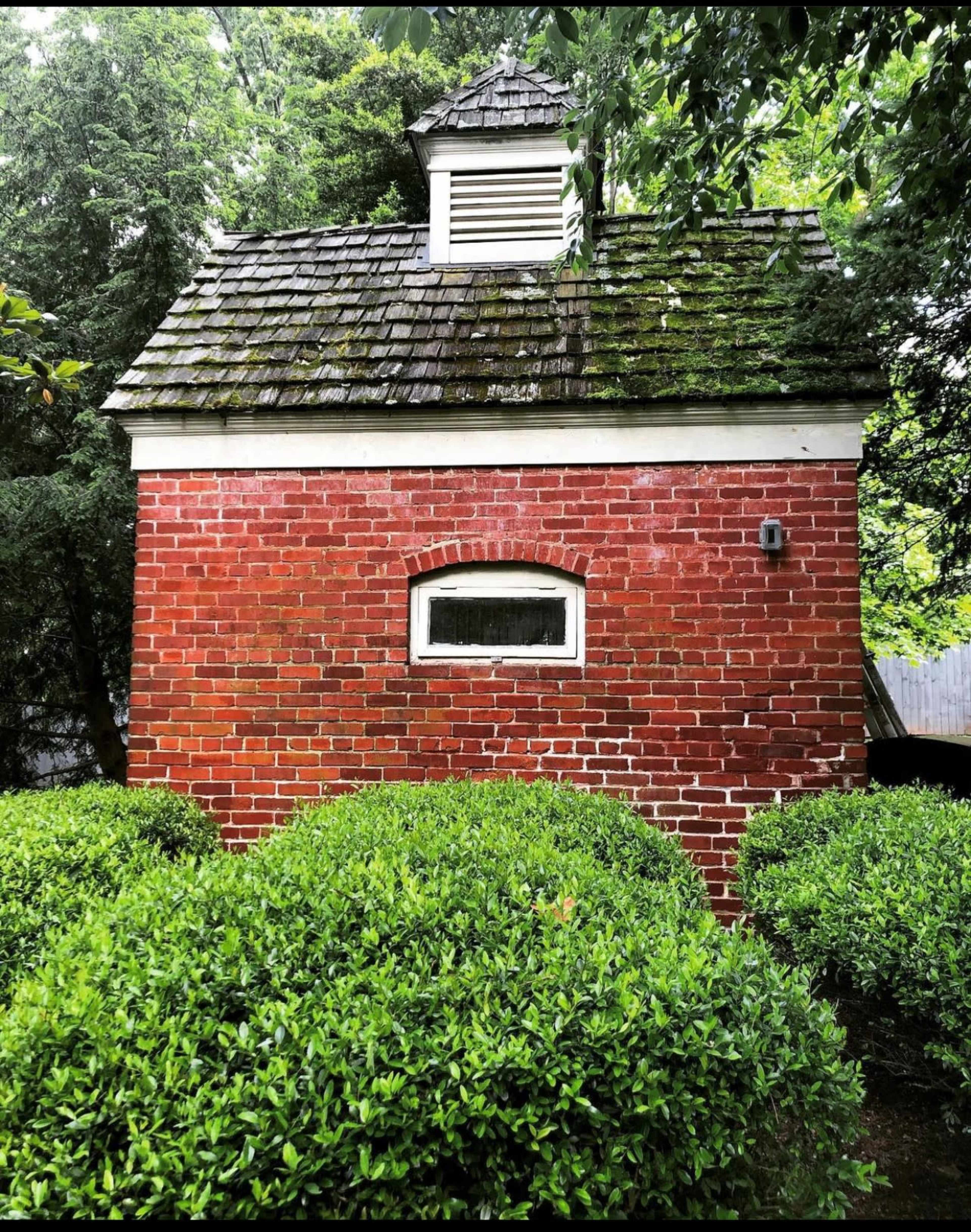 The image shows a small red brick building with a sloped roof and an attic vent, surrounded by neatly trimmed green bushes.