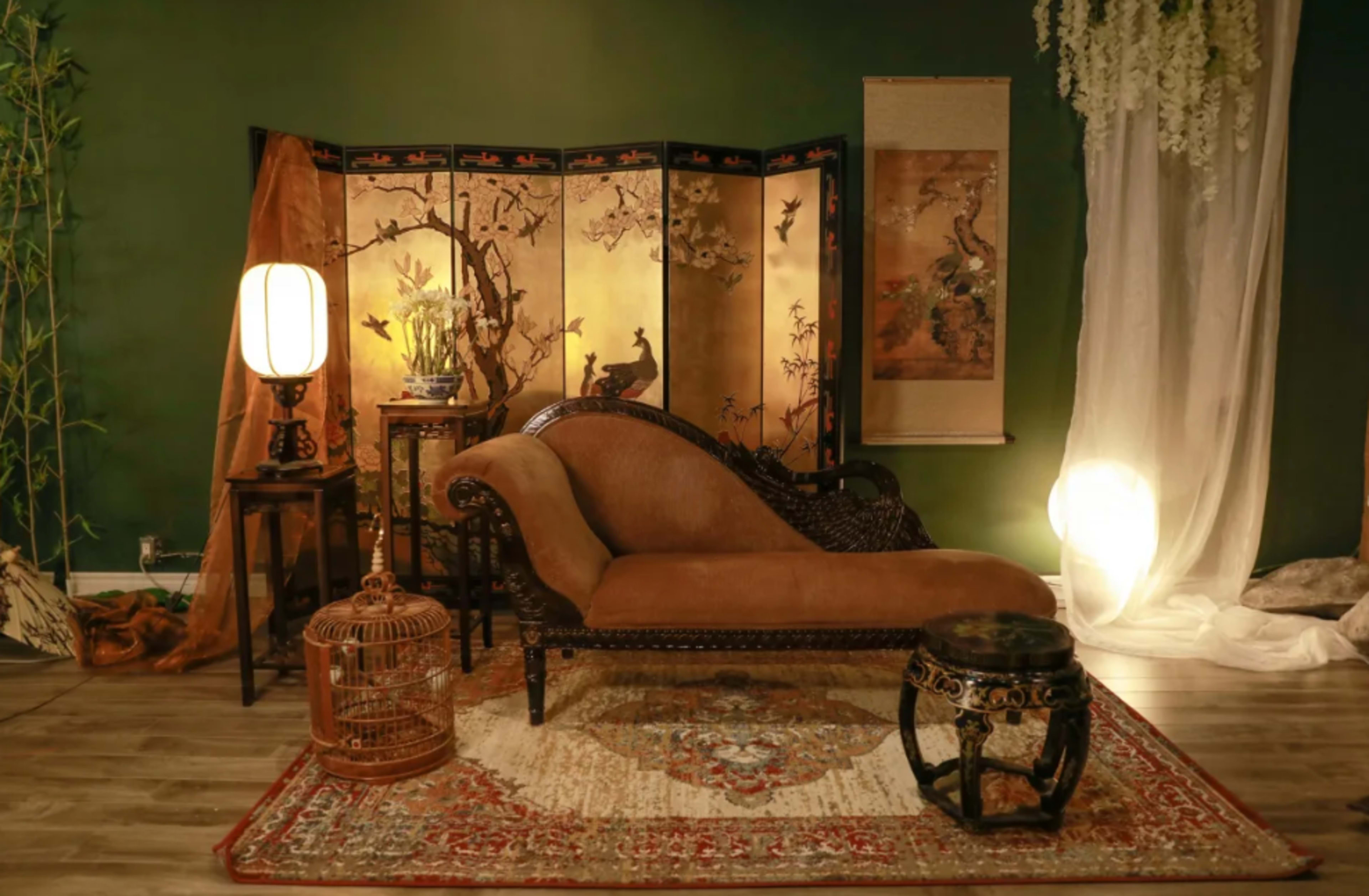 An elegantly furnished room featuring a vintage chaise lounge, decorative screens, and various ornamental pieces against a dark green wall.