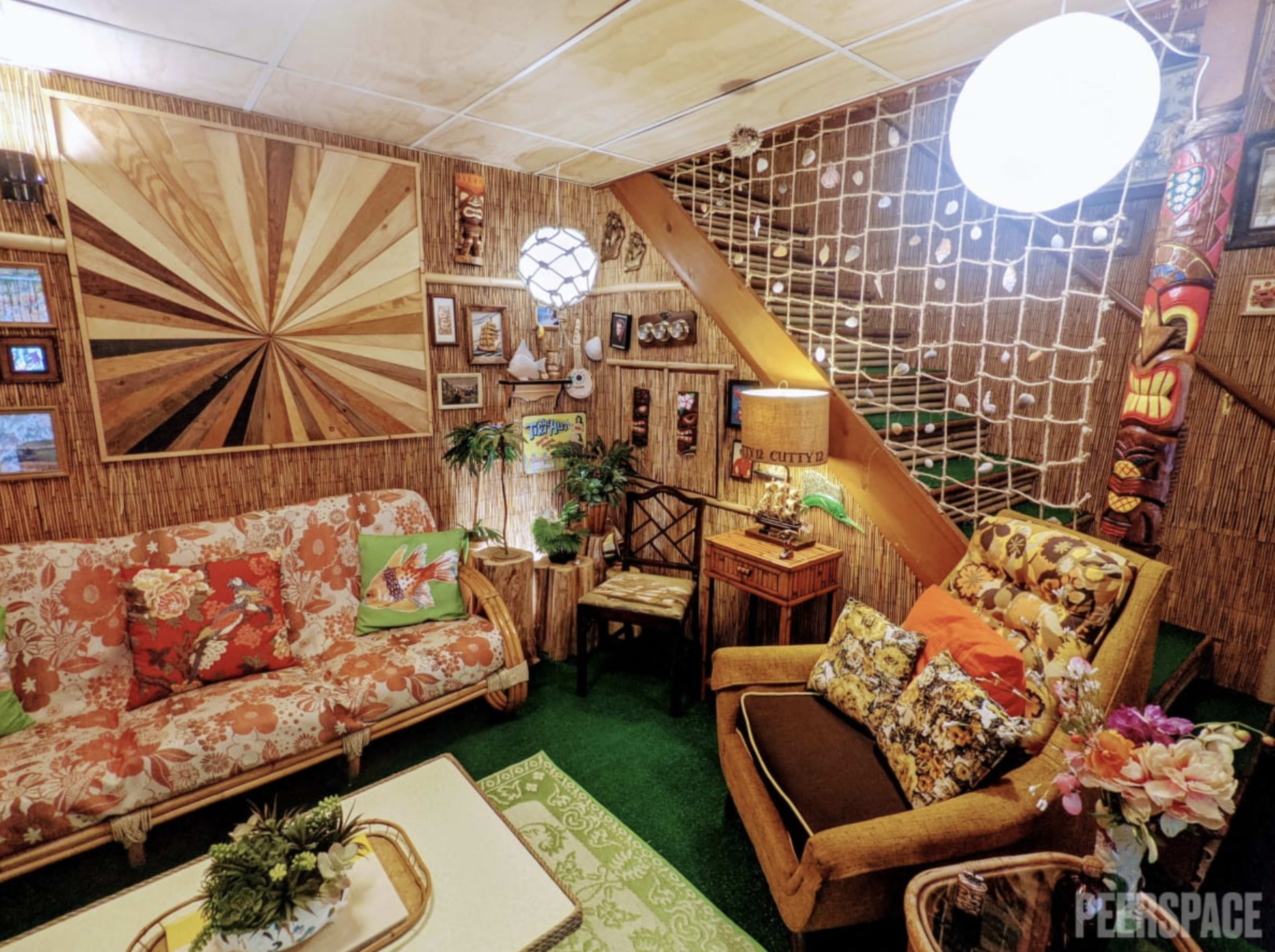 The image shows a cozy living room with bamboo walls, vintage furniture, and a green carpet, featuring floral-patterned sofas and various decorative items on display.