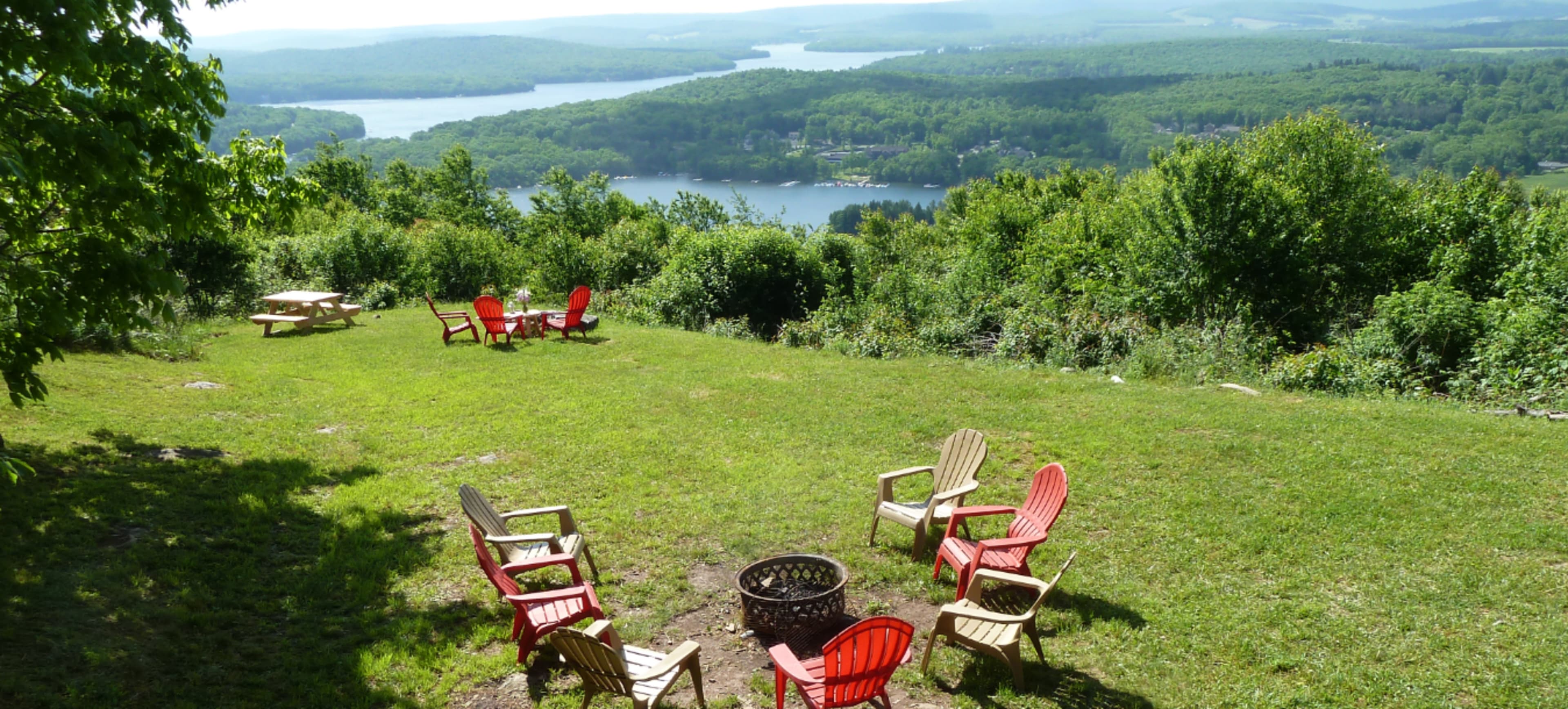 The image shows a grassy area overlooking a lake, featuring a circle of wooden chairs around a fire pit with additional seating in the background.