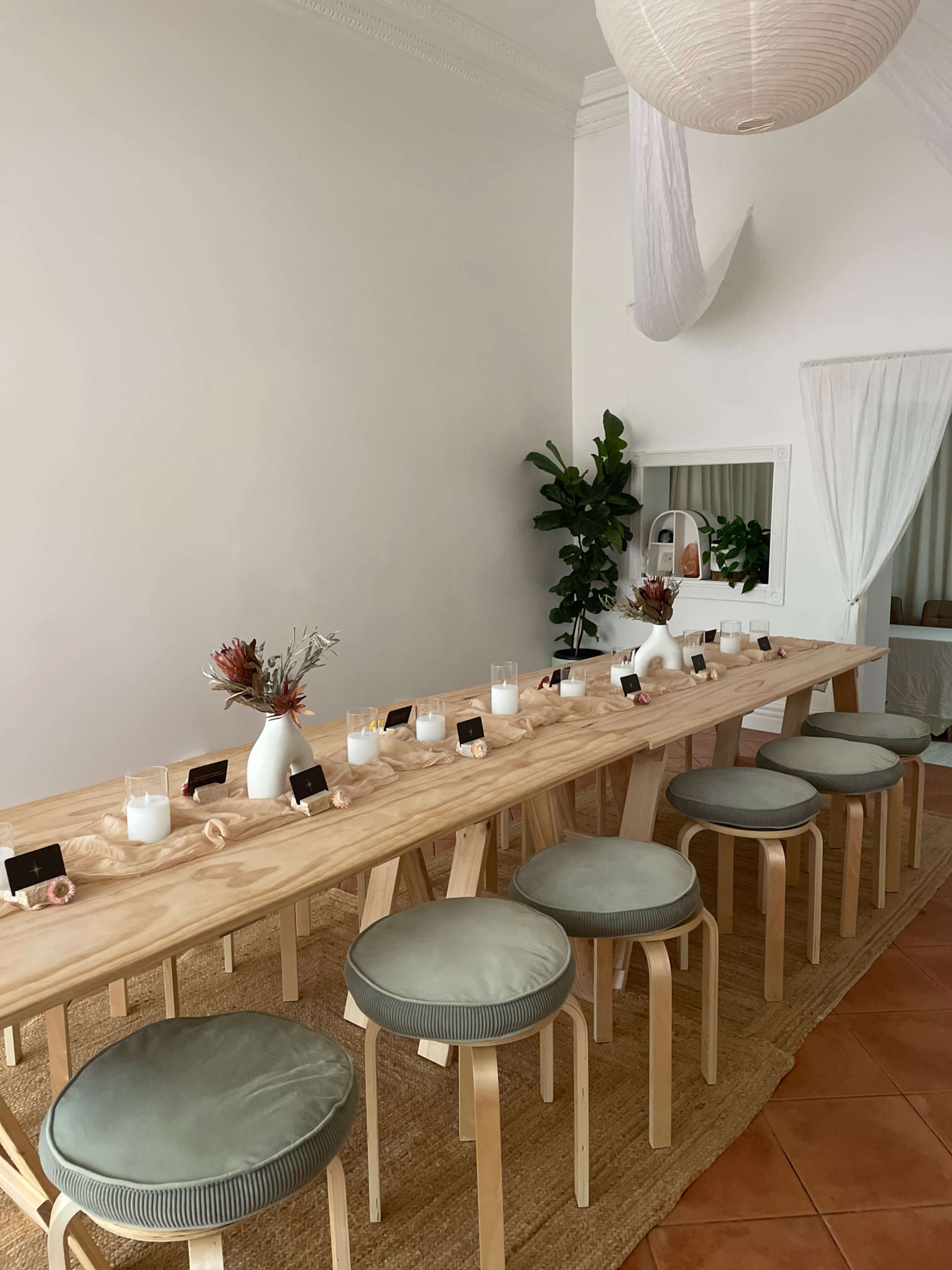 A long wooden table is set with white vases and candles, surrounded by light-colored stools, in a bright, minimalistic room with a potted plant.