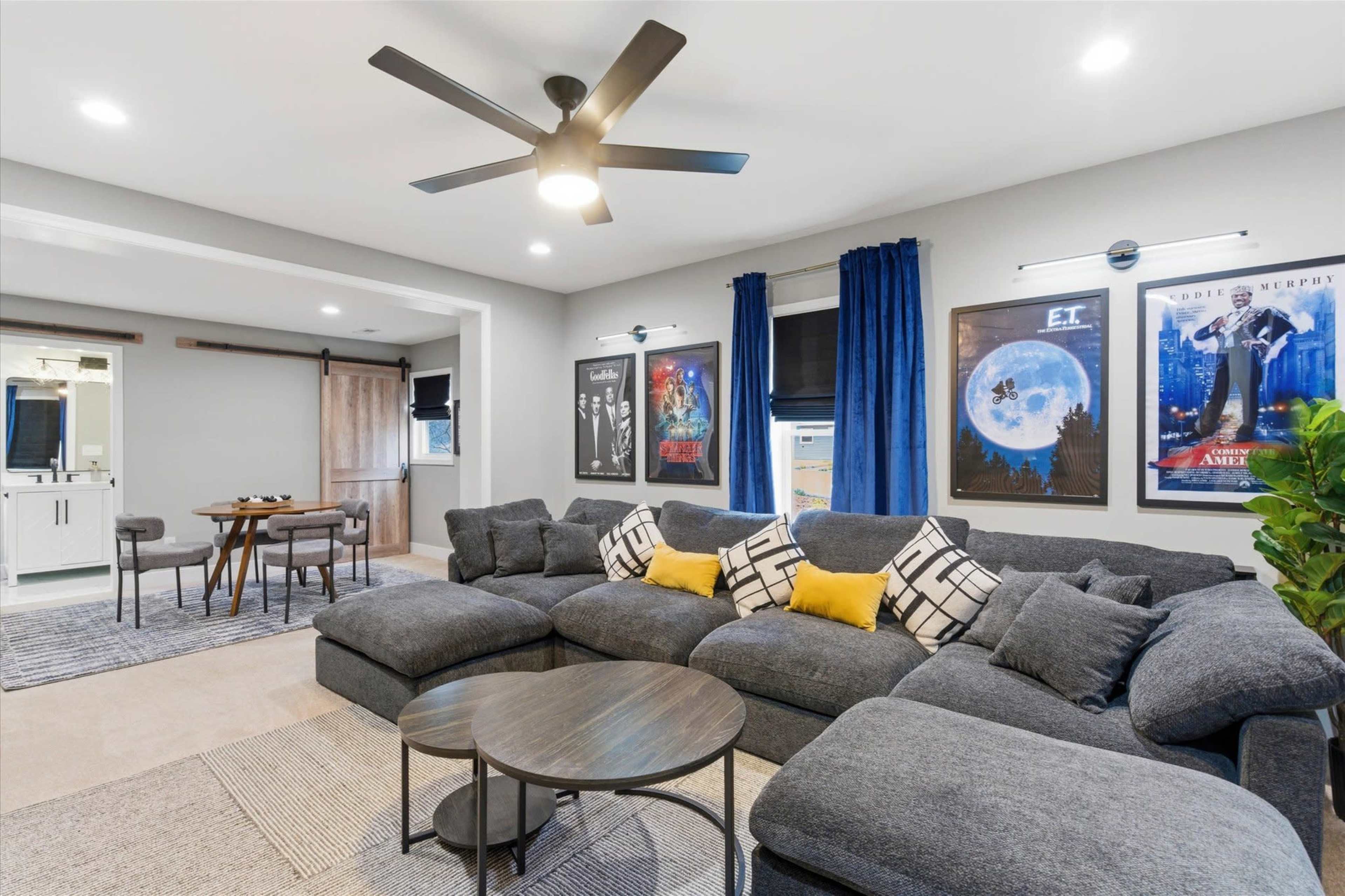 The image shows a modern living room featuring a large sectional sofa, a round coffee table, and several framed movie posters on the walls.