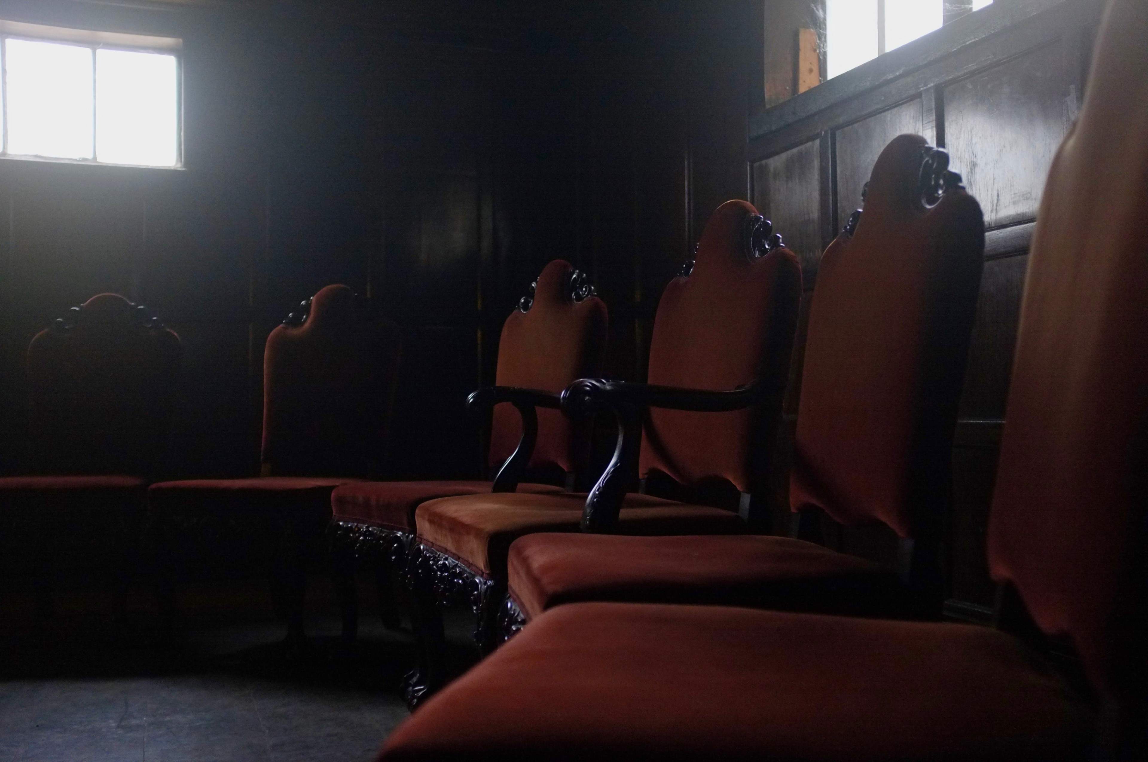 A row of ornate, upholstered chairs is positioned in a dimly lit room with a small window.