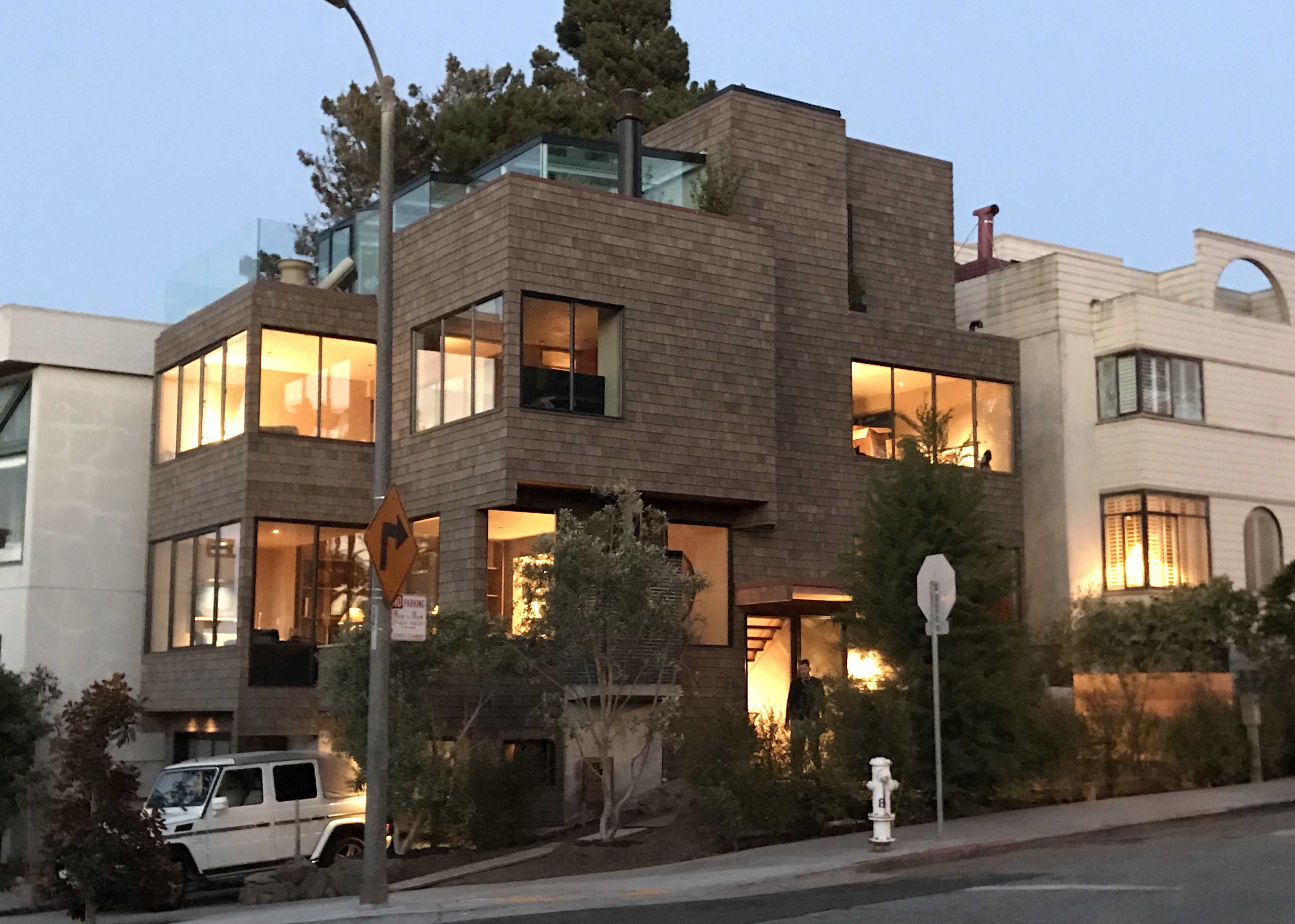 The image shows a modern multi-story house with large windows and a wooden exterior, situated on a street corner.