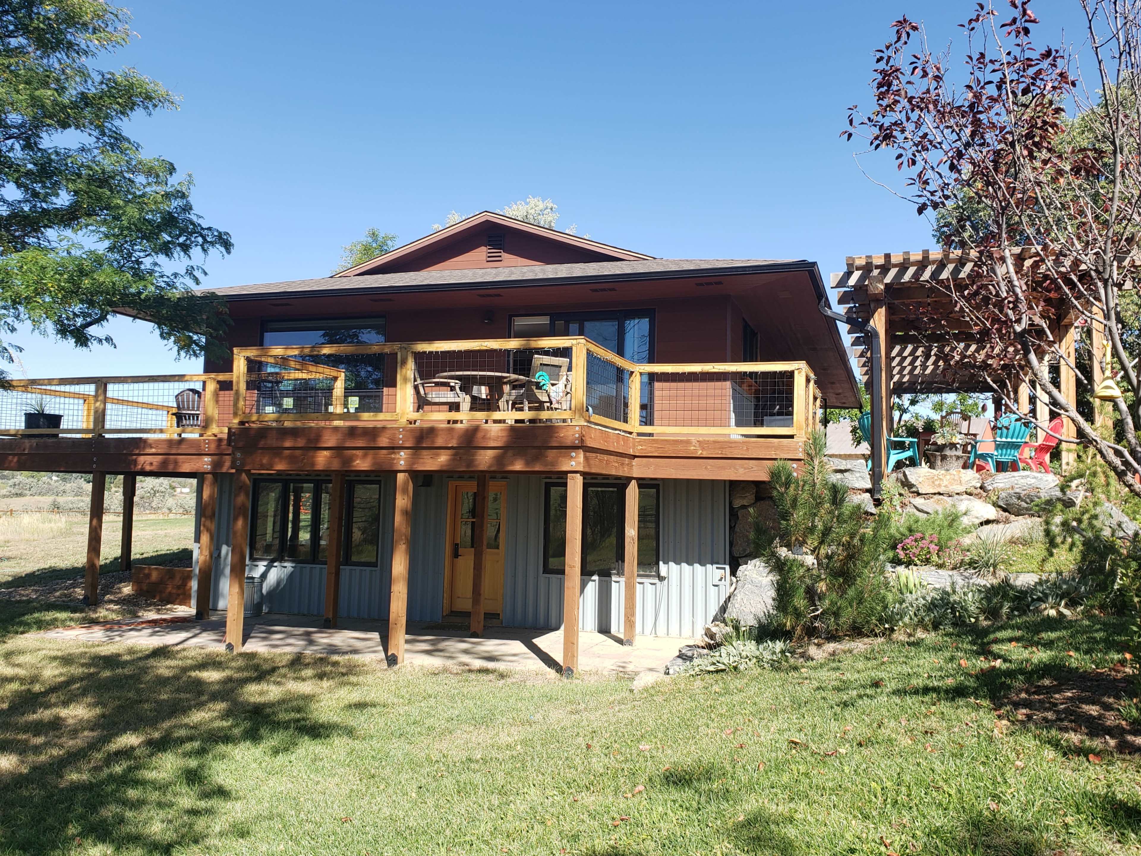The image shows a two-story house with a wooden deck, surrounded by greenery and a pergola with outdoor seating in the background.