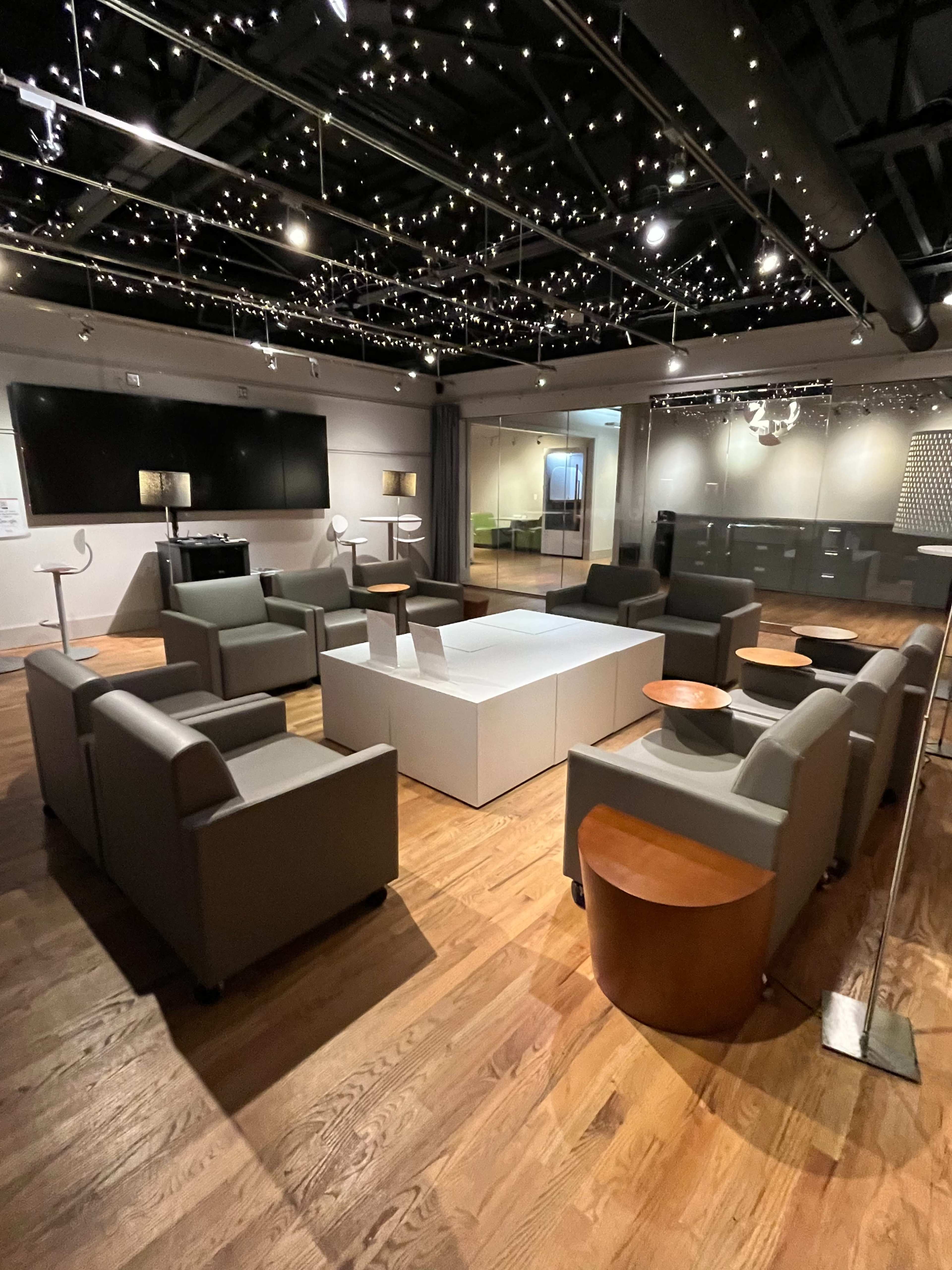 The image shows a modern lounge area with beige sofas arranged around a white coffee table, illuminated by string lights overhead.