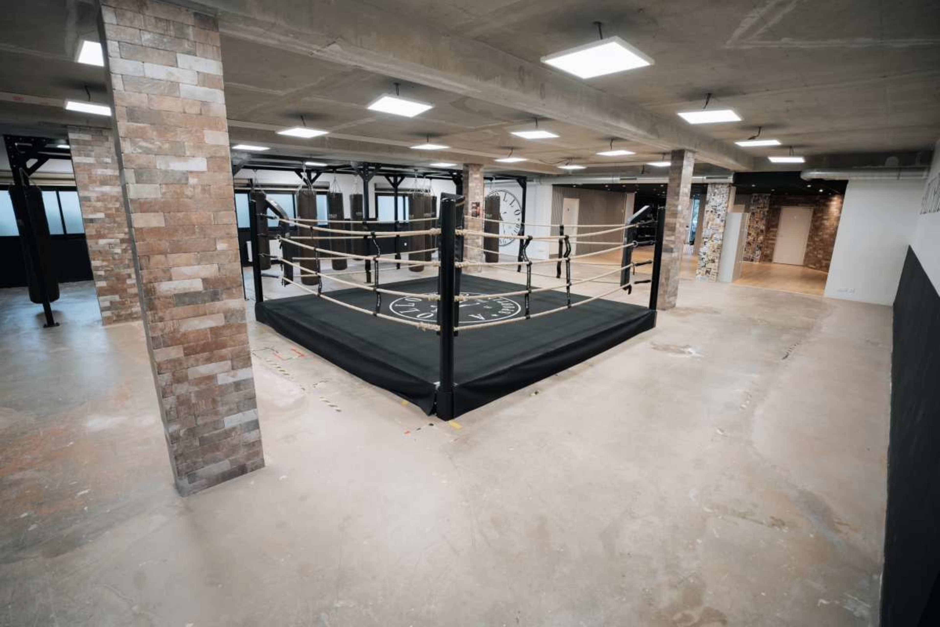 The image shows an empty boxing ring set up in a spacious gym with exposed brick walls and concrete flooring.