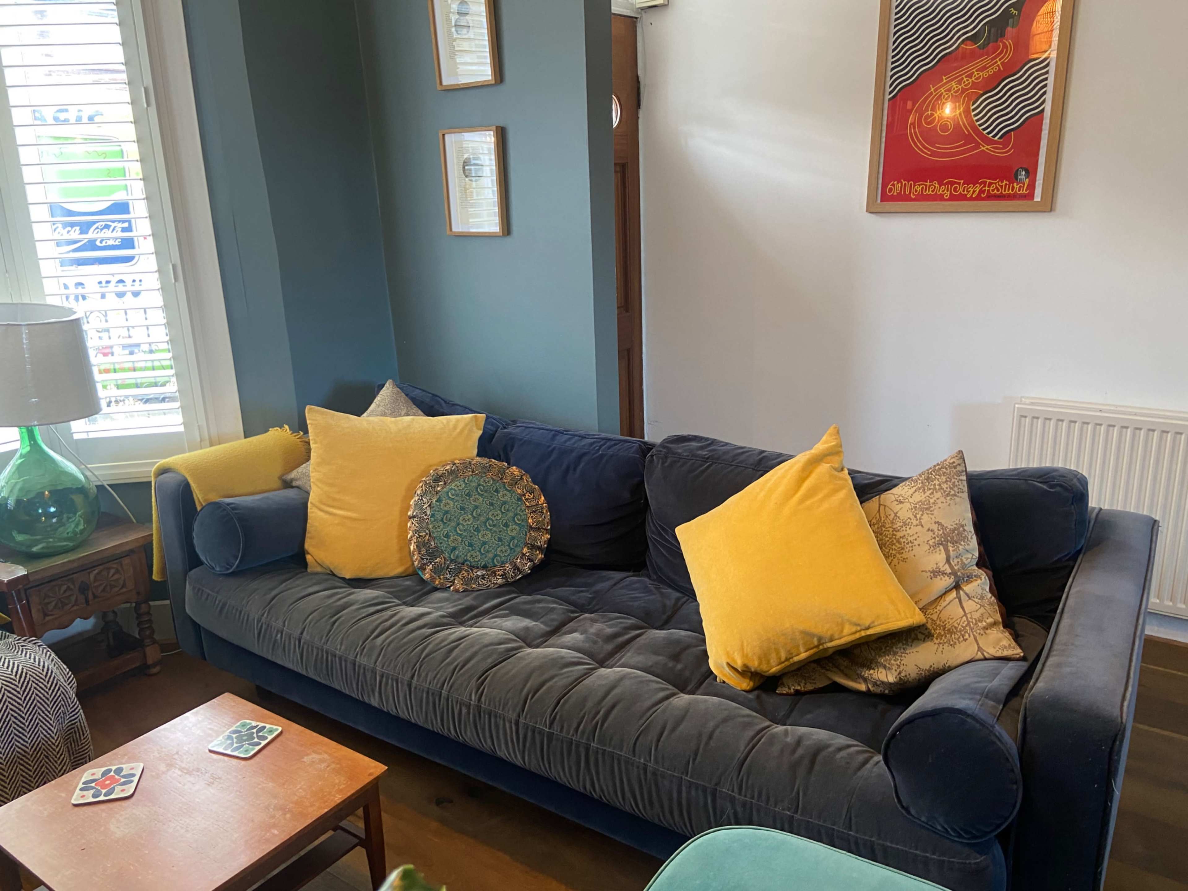 A blue velvet sofa with yellow cushions is placed next to a wooden table in a brightly lit room.