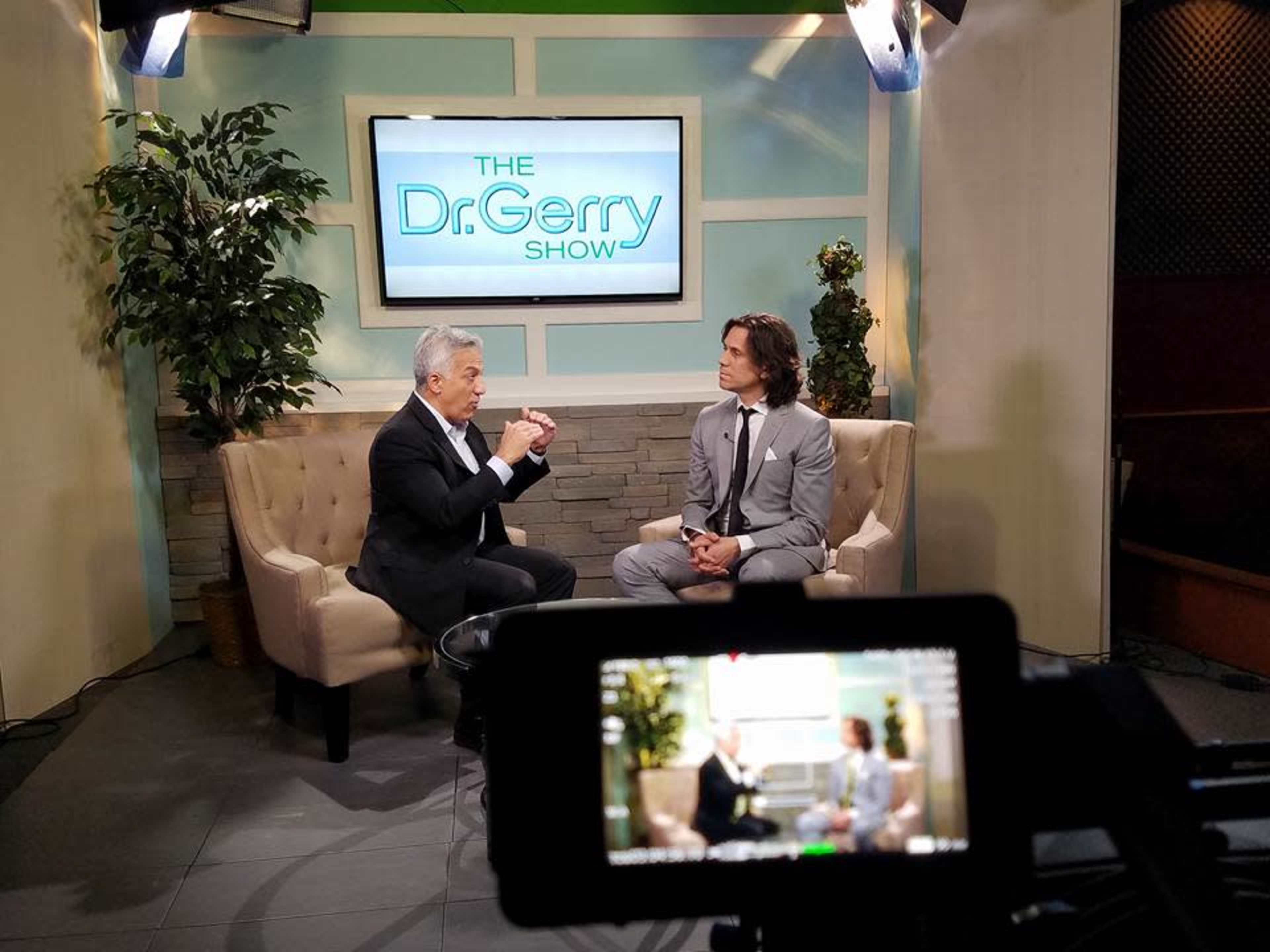 The image shows two men seated on a couch in a television studio, with the set displaying "The Dr. Gerry Show" on a screen behind them.