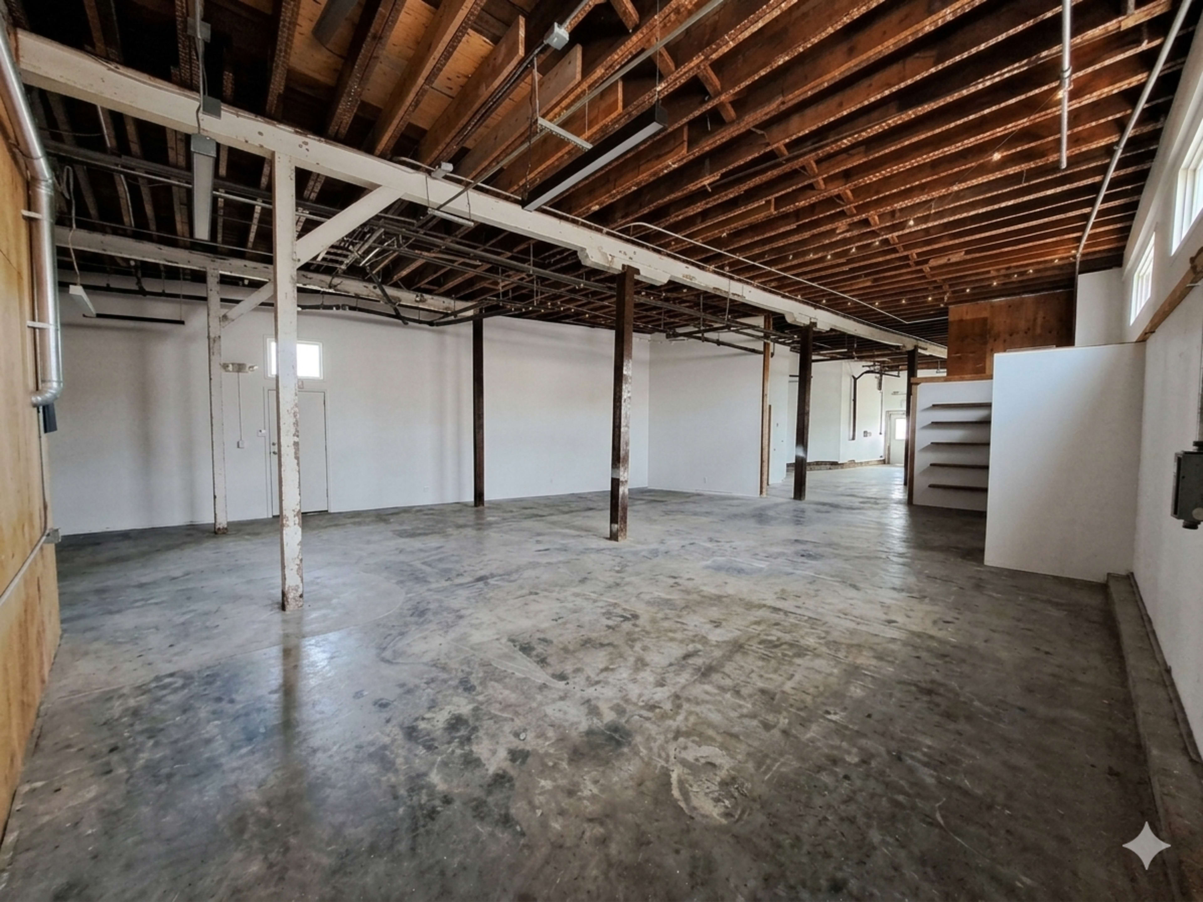The image shows a spacious, empty interior area with exposed beams and a concrete floor, featuring white walls and minimal fixtures.