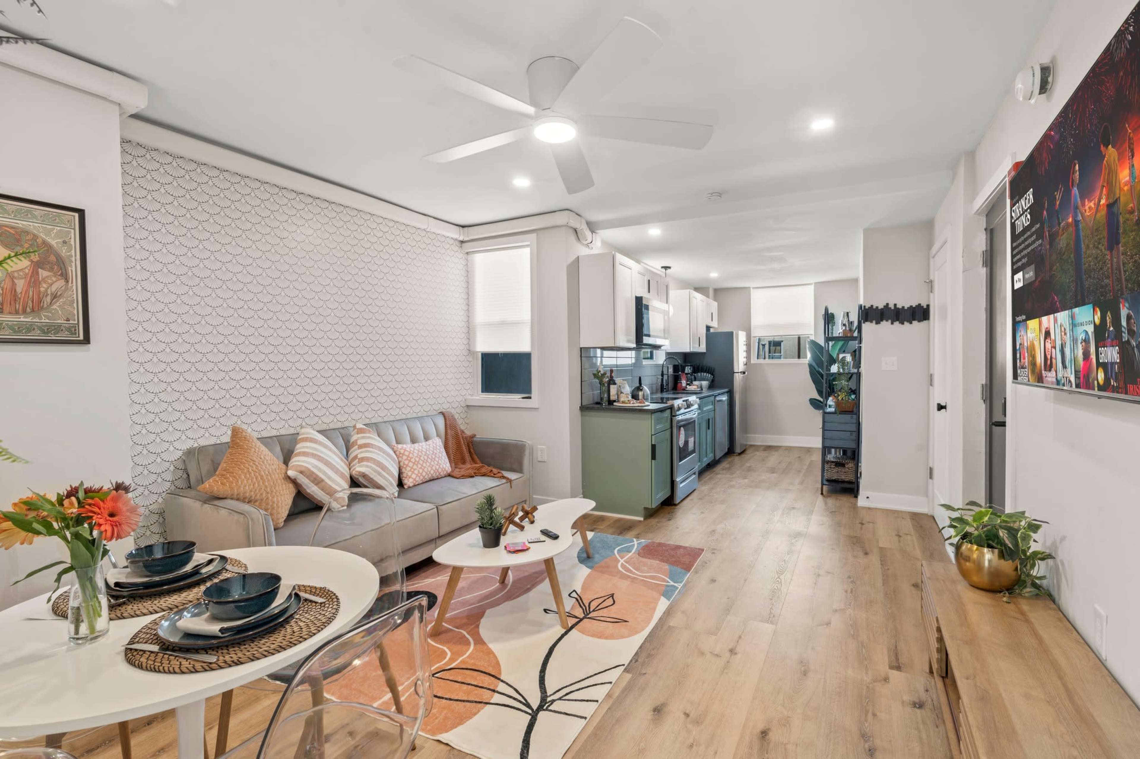 The image shows a modern living space featuring a light gray sofa, a dining table set for two, and an open kitchen area with wooden flooring.