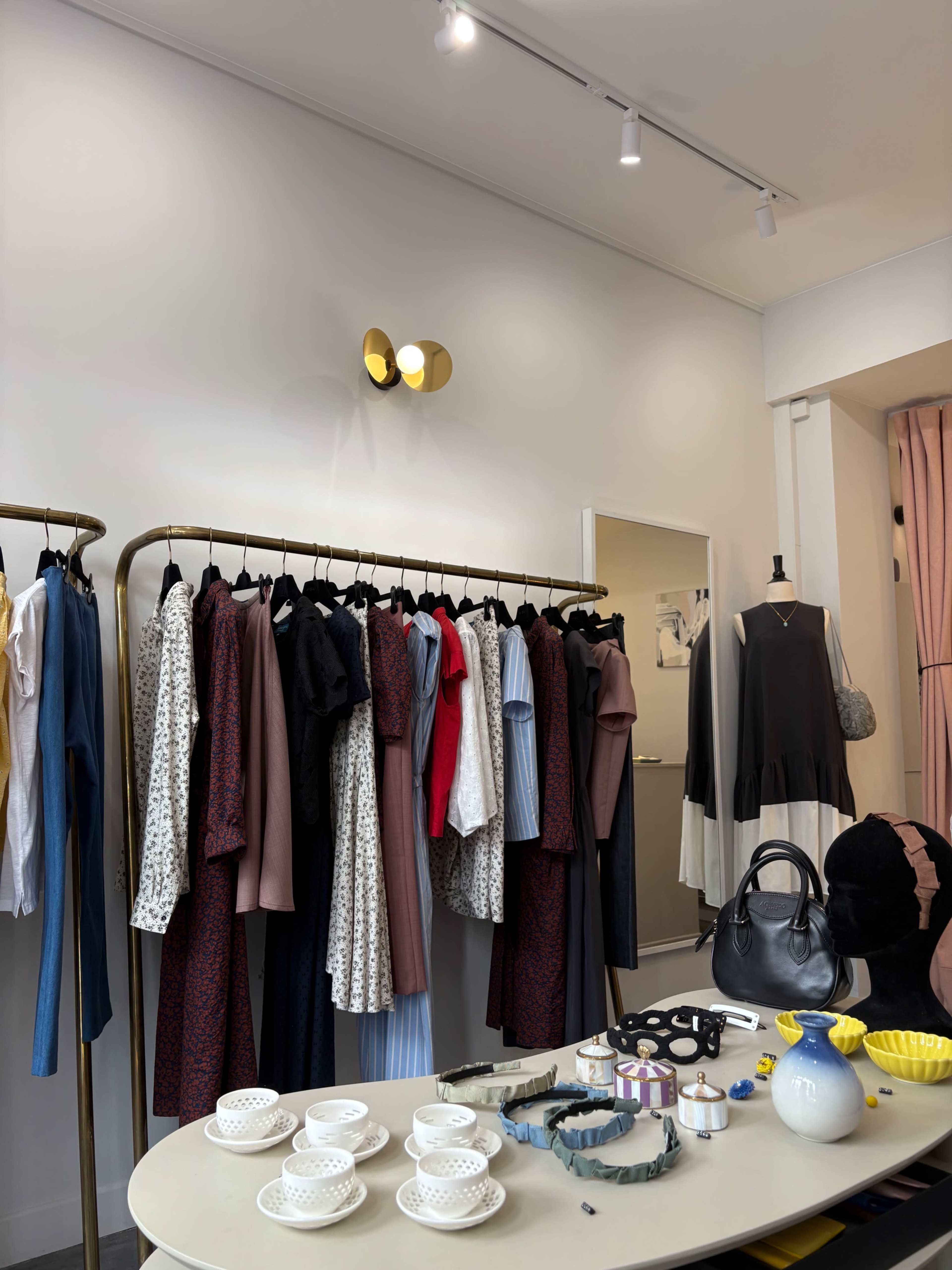 A clothing rack displays a variety of garments, while a table showcases several decorative items and dishware in a boutique setting.