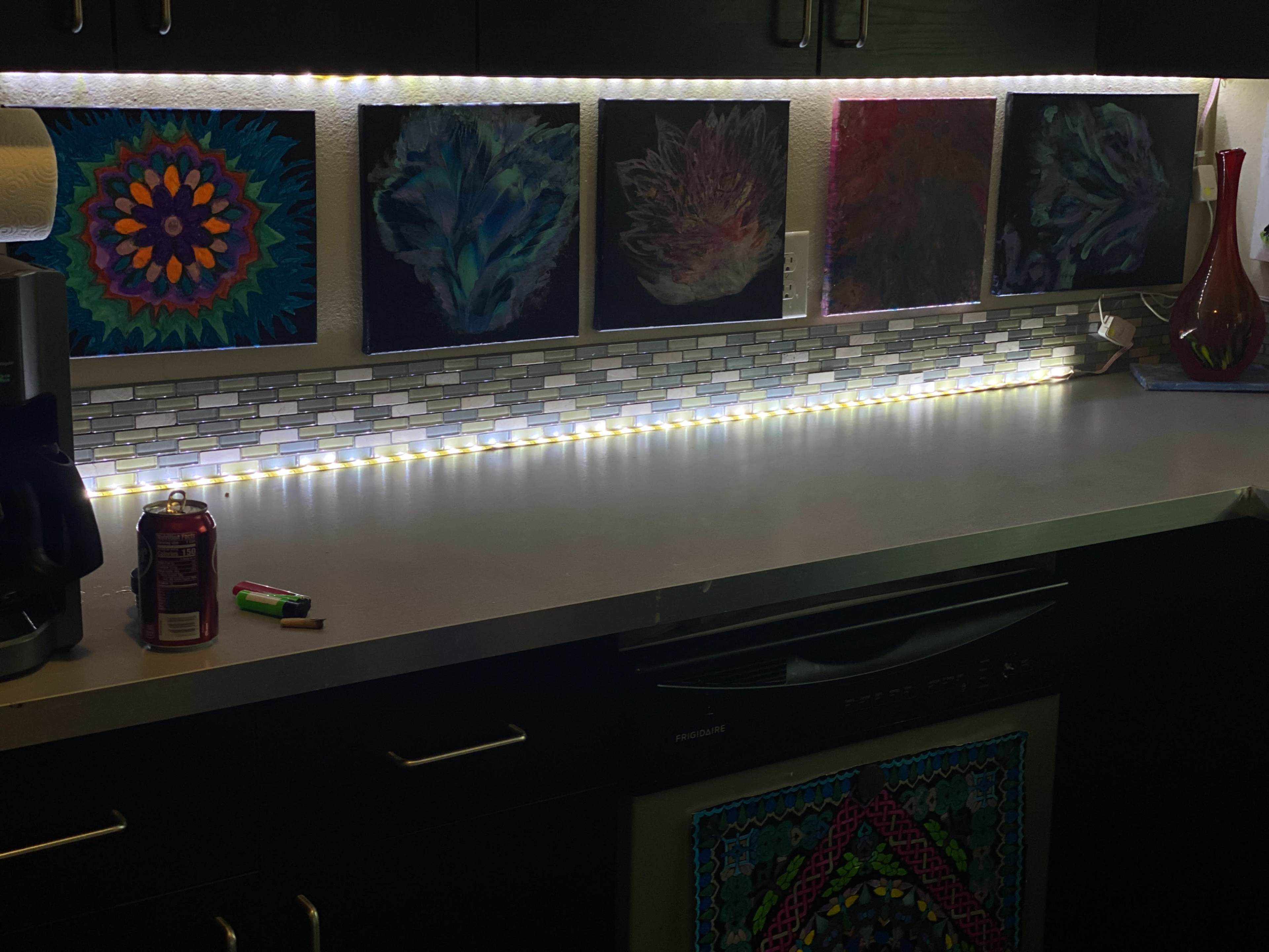 The image shows a kitchen counter with illuminated artwork above a backsplash, complemented by a coffee maker and a soft drink can on the surface.