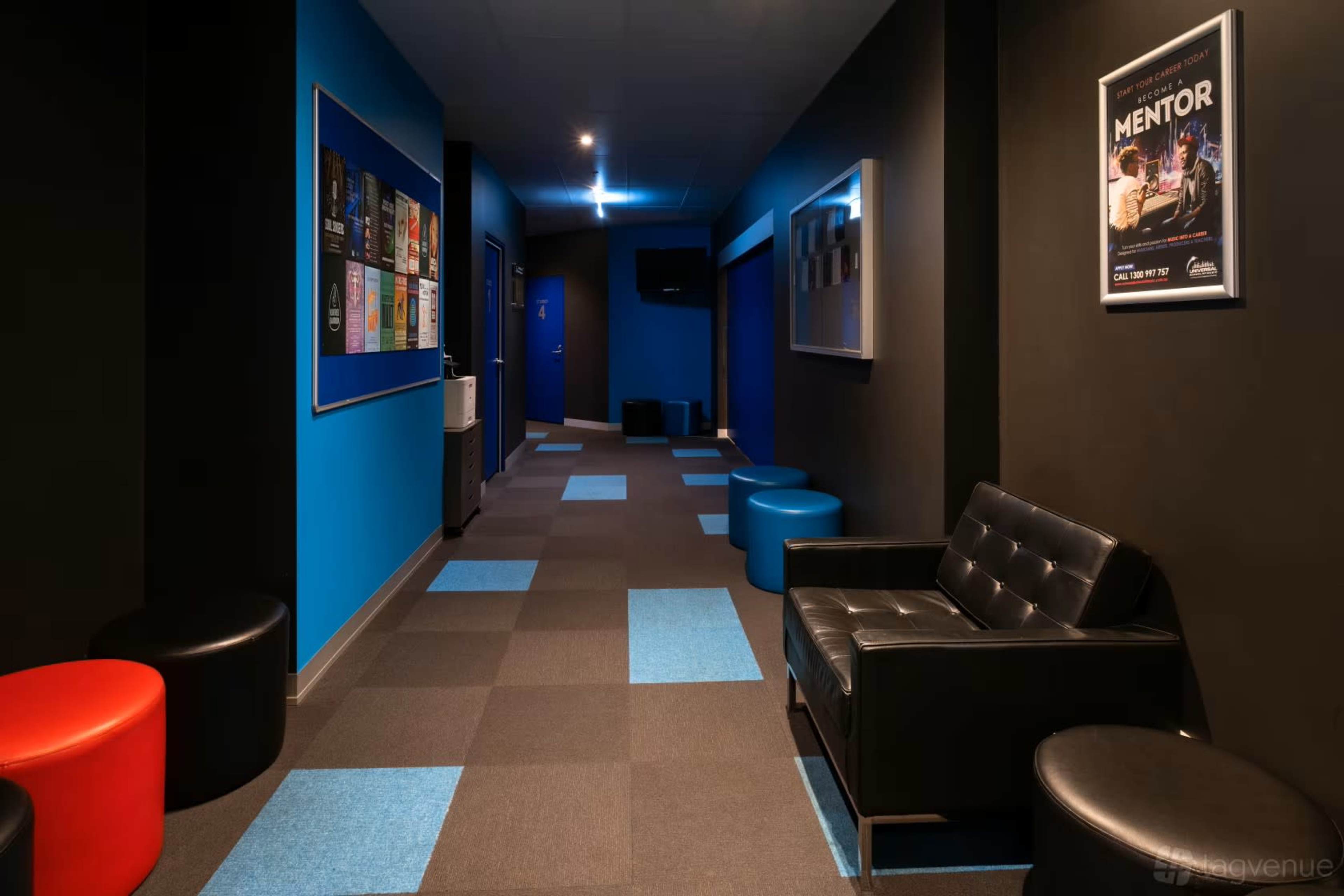 The image shows a modern hallway with black and blue walls, featuring a mix of circular red and blue stools, a black couch, and a bulletin board displaying posters.