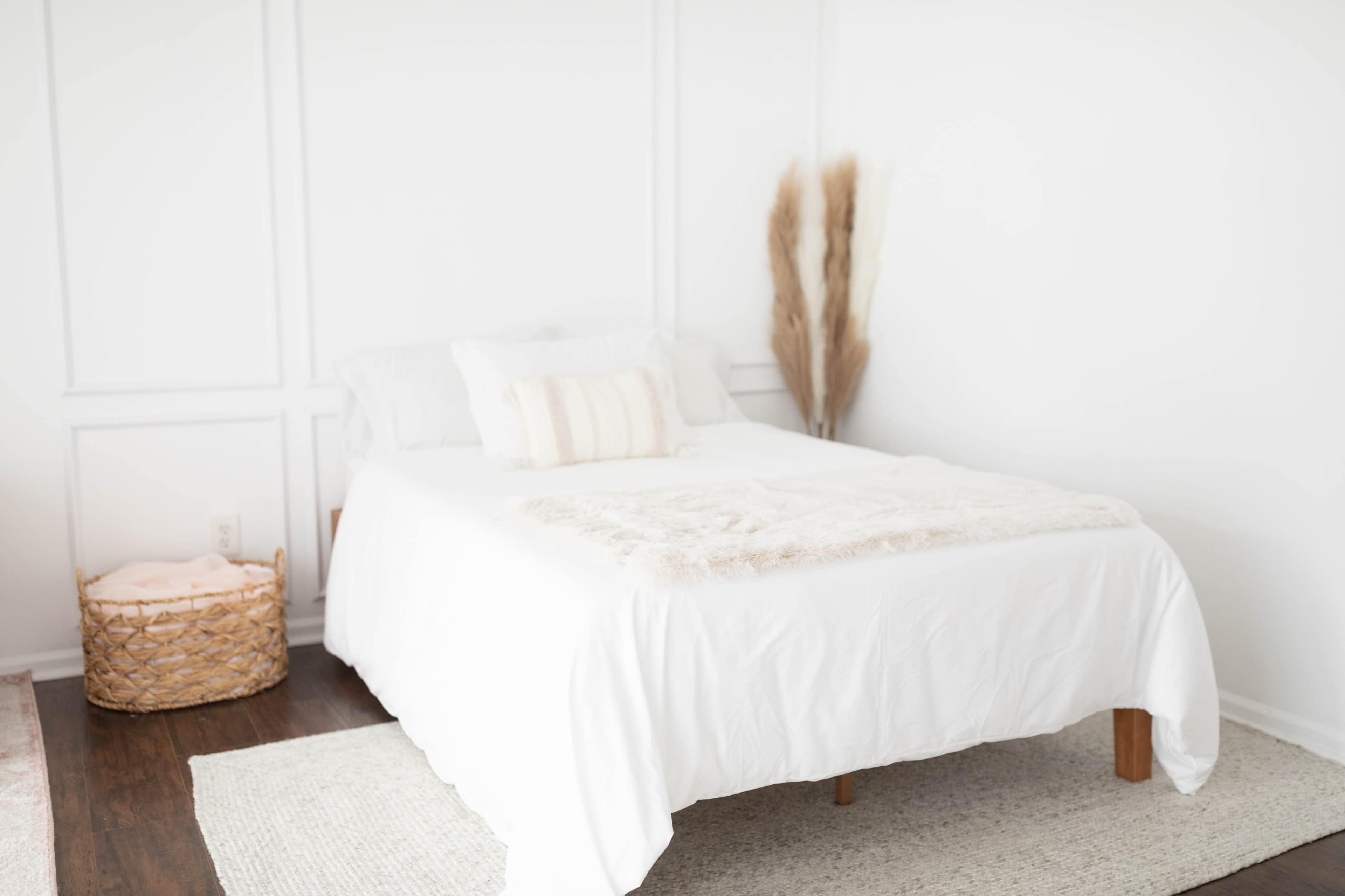 A neatly arranged bedroom features a white bed with a light coverlet, a woven basket in the corner, and decorative dried plants against the wall.