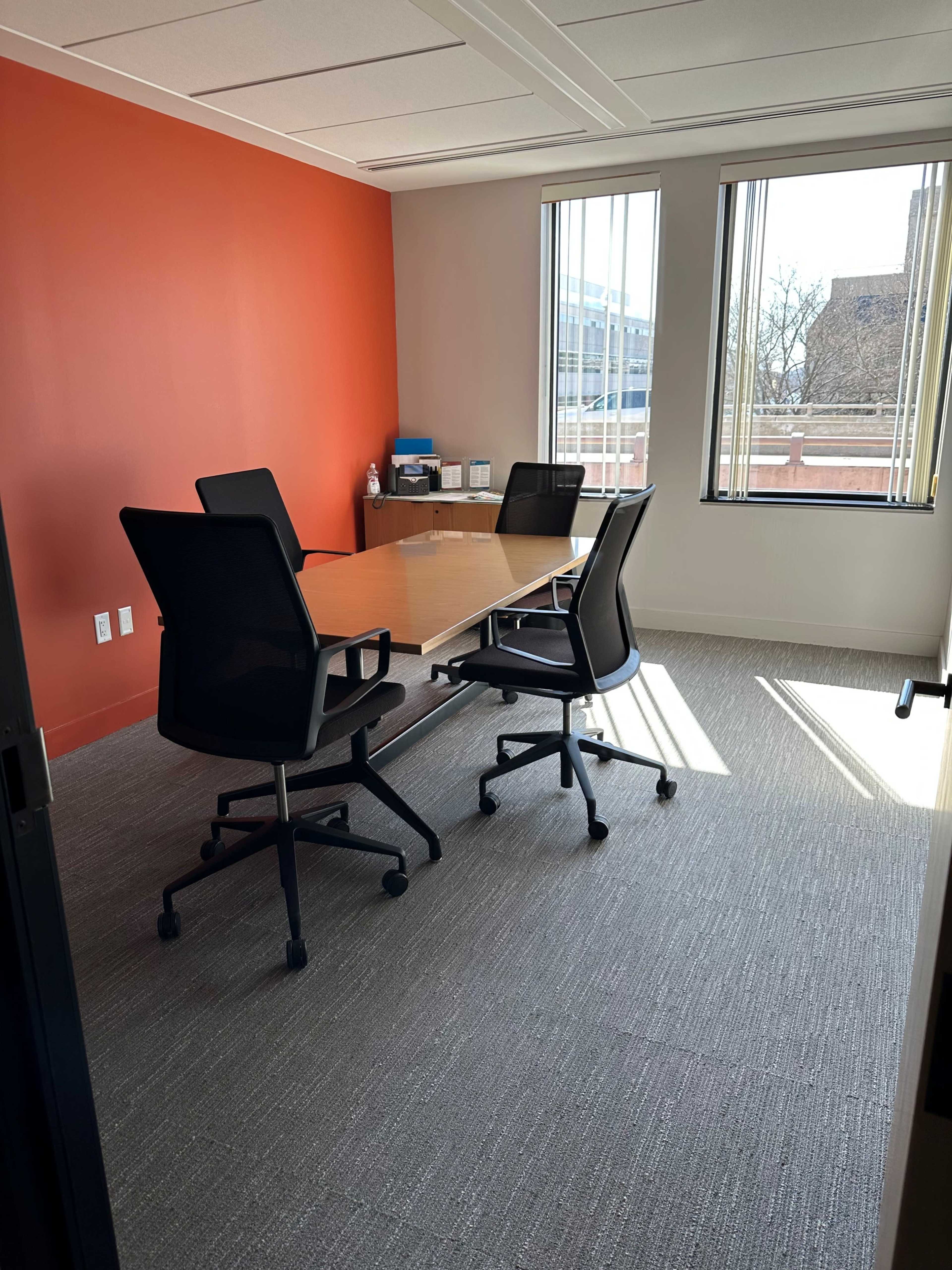 The image shows a bright, empty conference room with a wooden table and four black chairs, set against an orange accent wall and large windows letting in natural light.