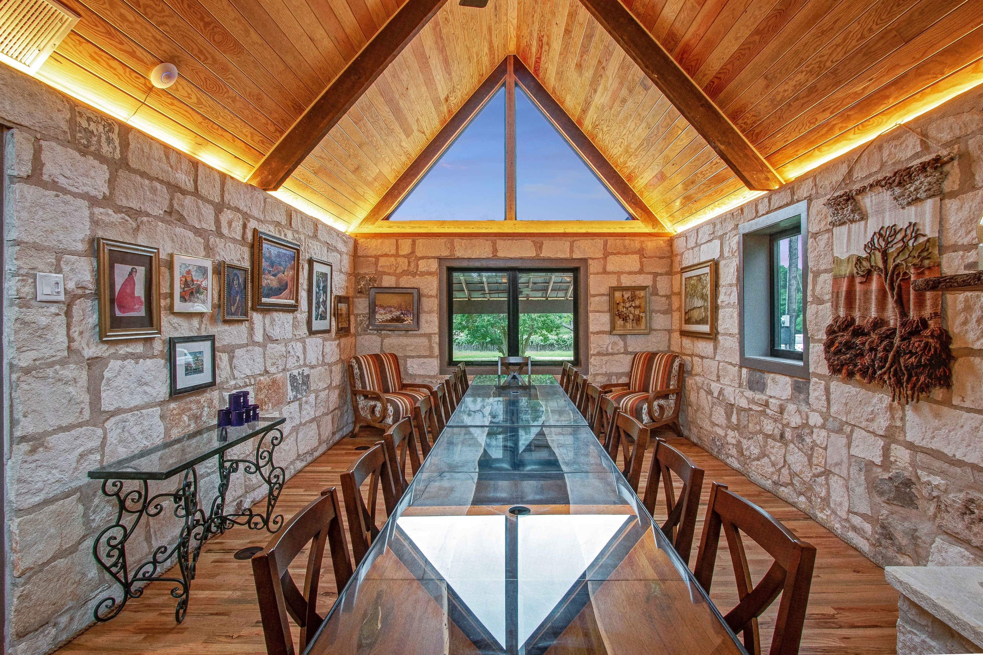 A modern dining room features a glass table surrounded by wooden chairs, stone walls, and large windows under a vaulted wooden ceiling.