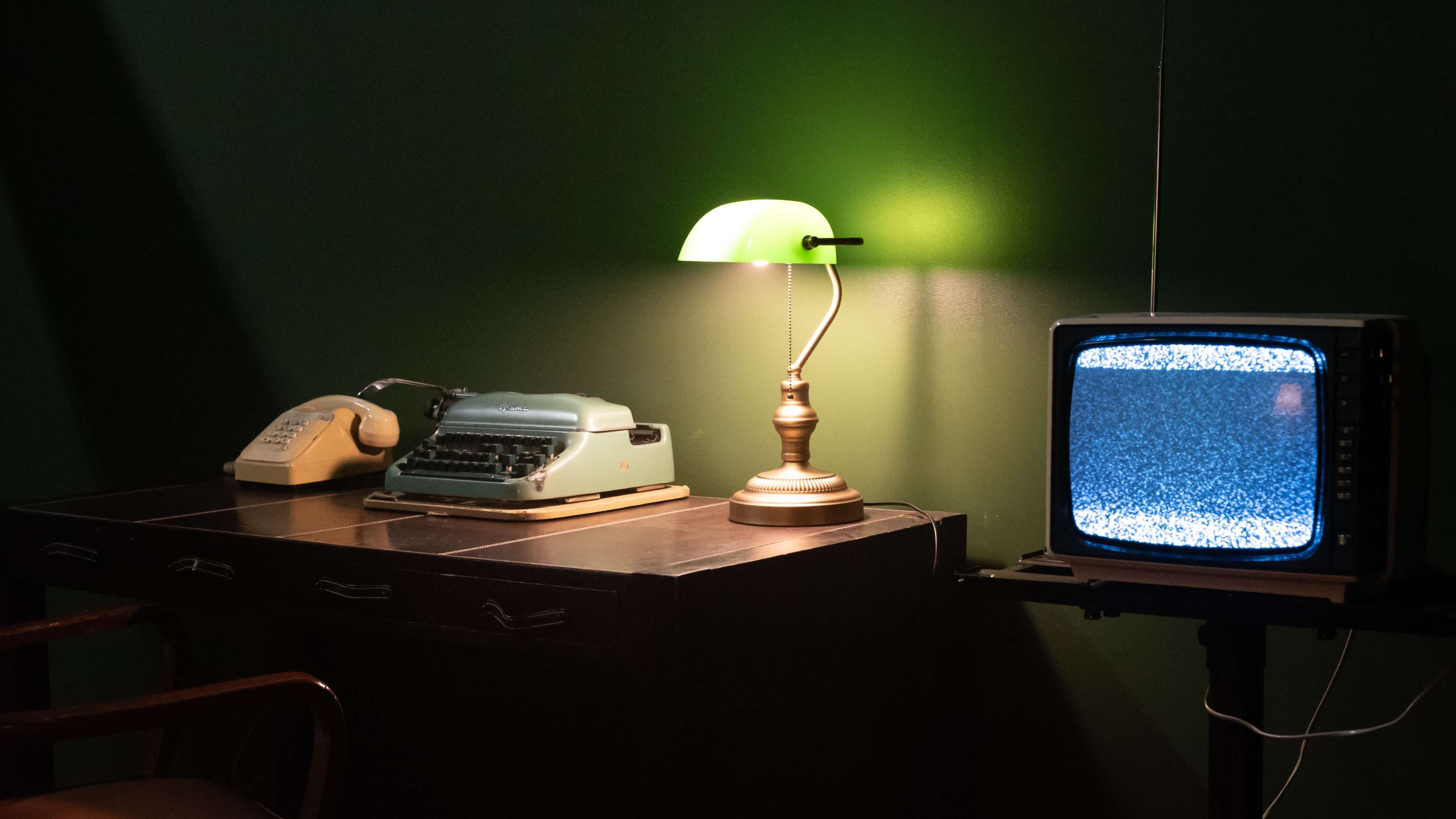 A vintage desk with a typewriter, a telephone, and a lamp sits next to an old television displaying static.