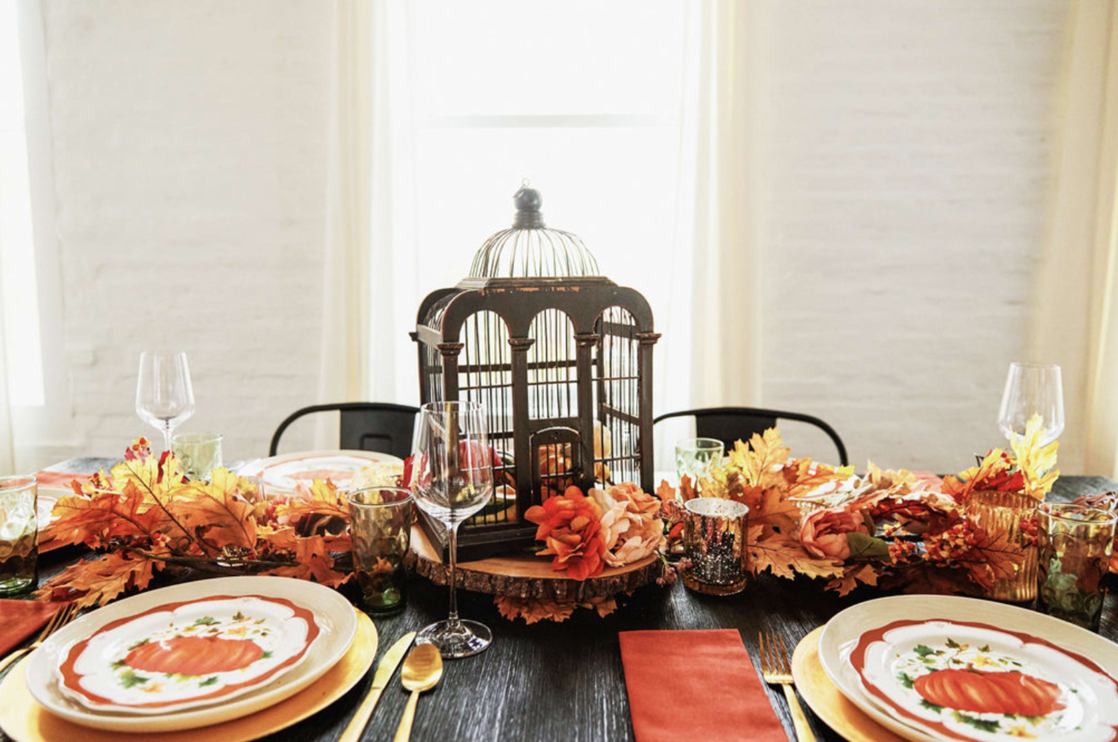 A beautifully arranged dining table is decorated with autumn leaves, pumpkin-themed plates, and a vintage birdcage centerpiece.