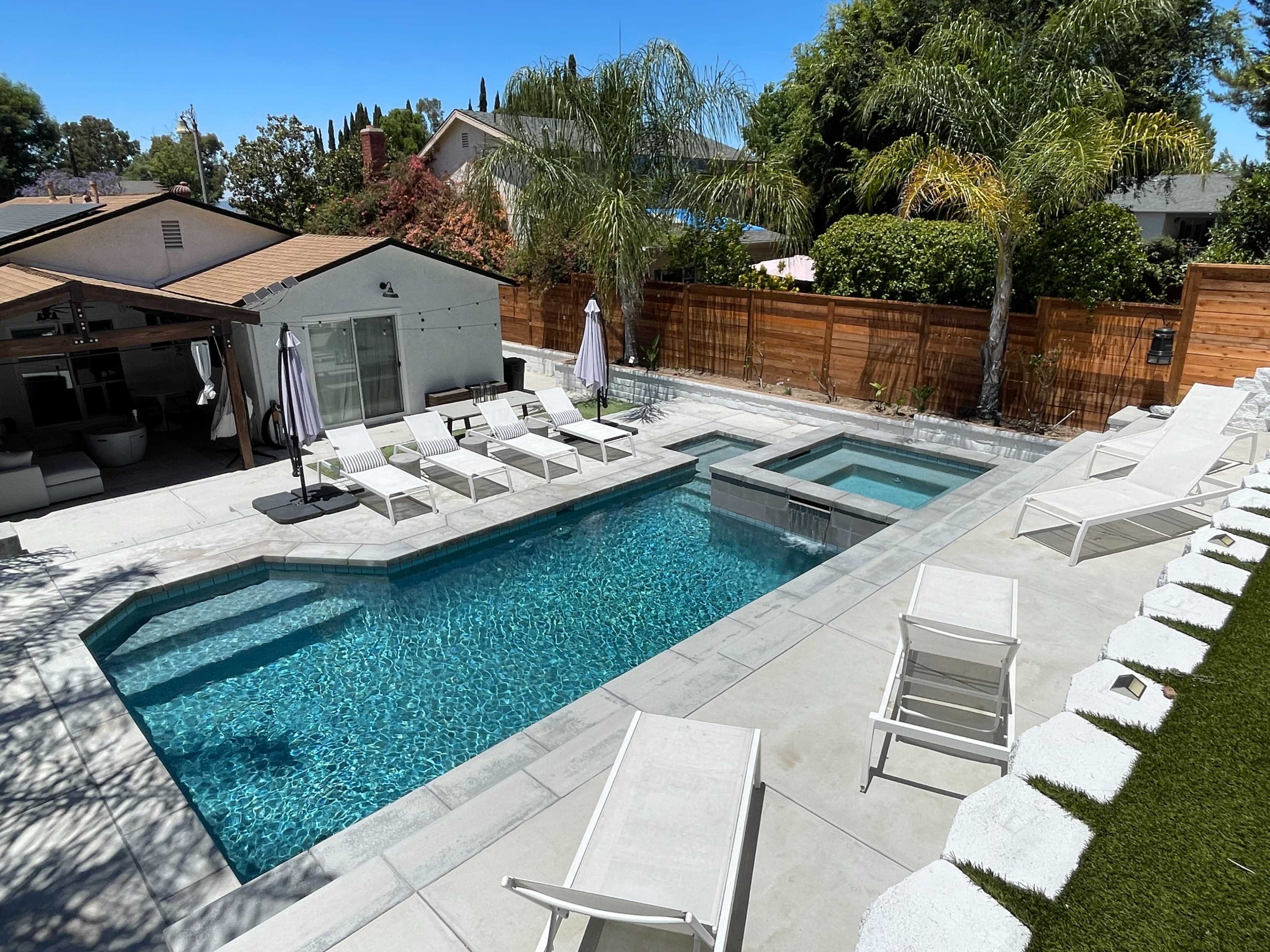 A backyard pool area featuring a rectangular swimming pool with a spa, surrounded by lounge chairs and palm trees.