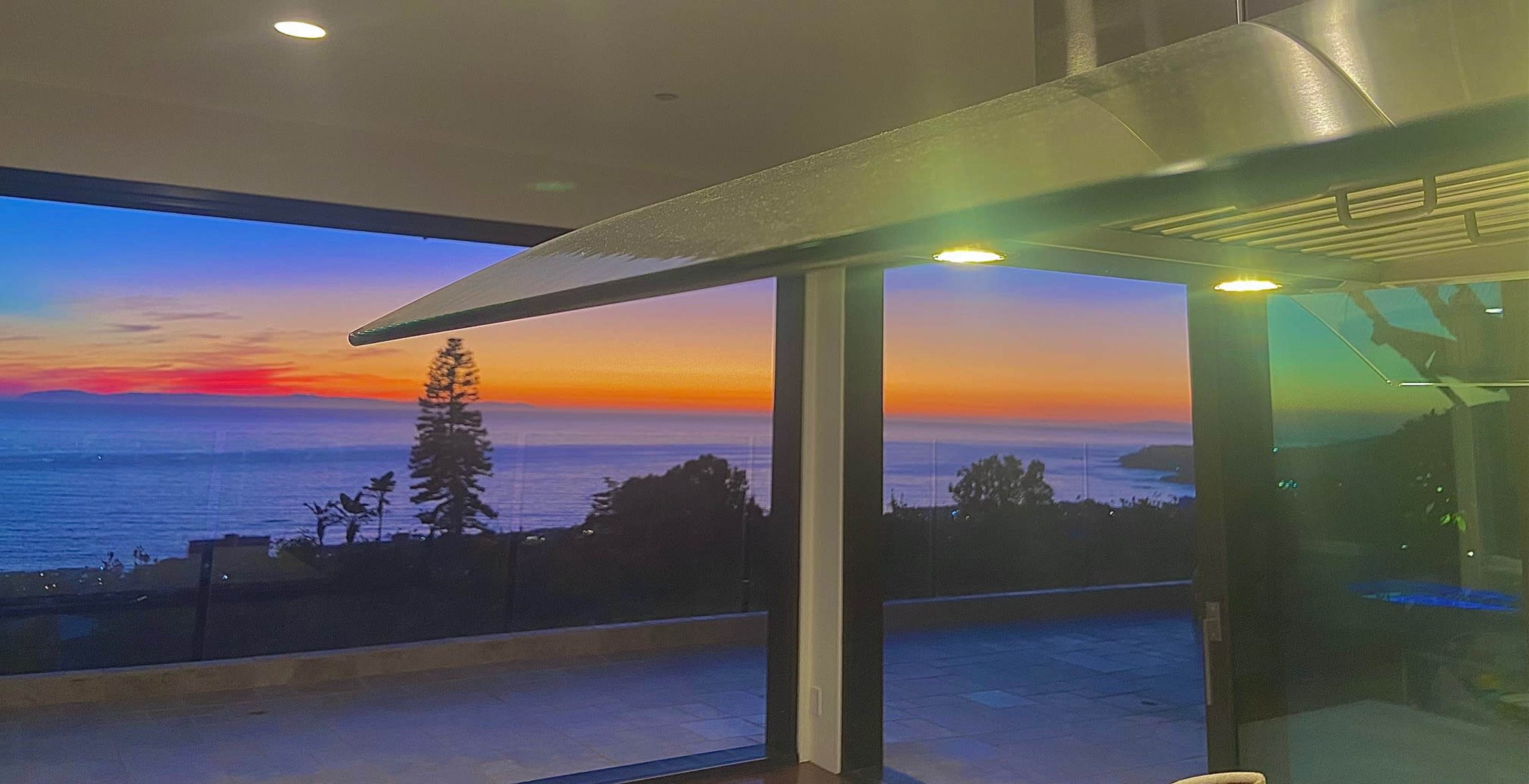 The image shows a coastal sunset visible through large windows, with a kitchen hood in the foreground.