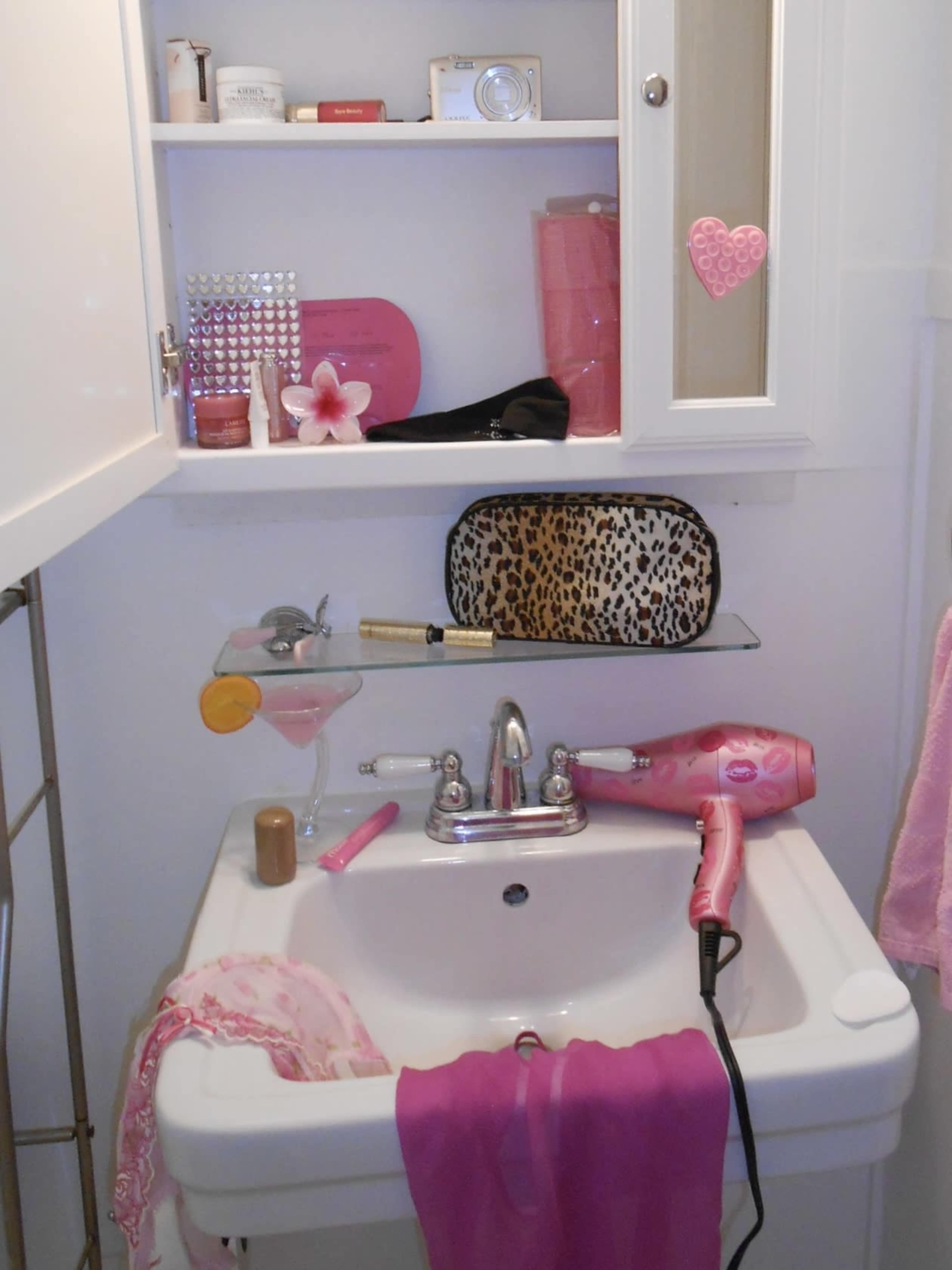 The image shows a small bathroom sink cluttered with various beauty products, a hairdryer, and a pink-colored towel.