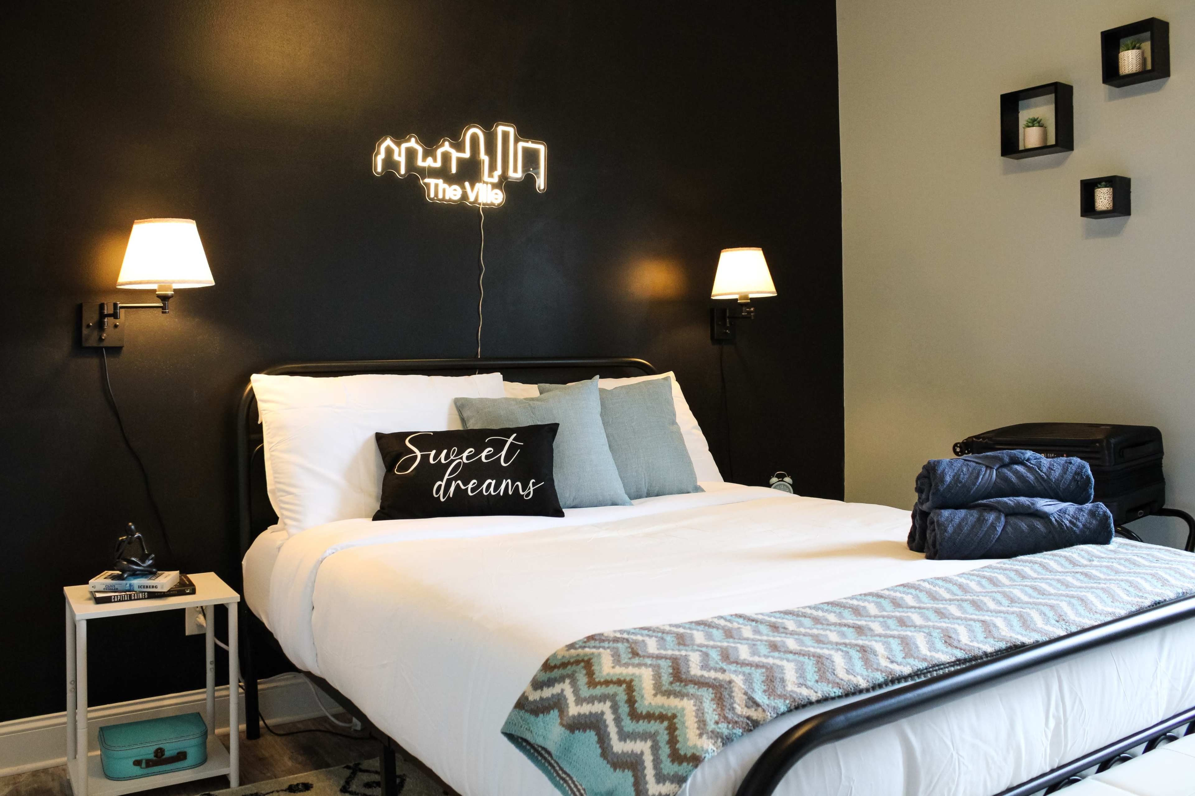 A neatly made bed with decorative pillows and a "Sweet dreams" throw pillow, situated in a room with dark walls, featuring wall sconces and a neon sign reading "The Ville."