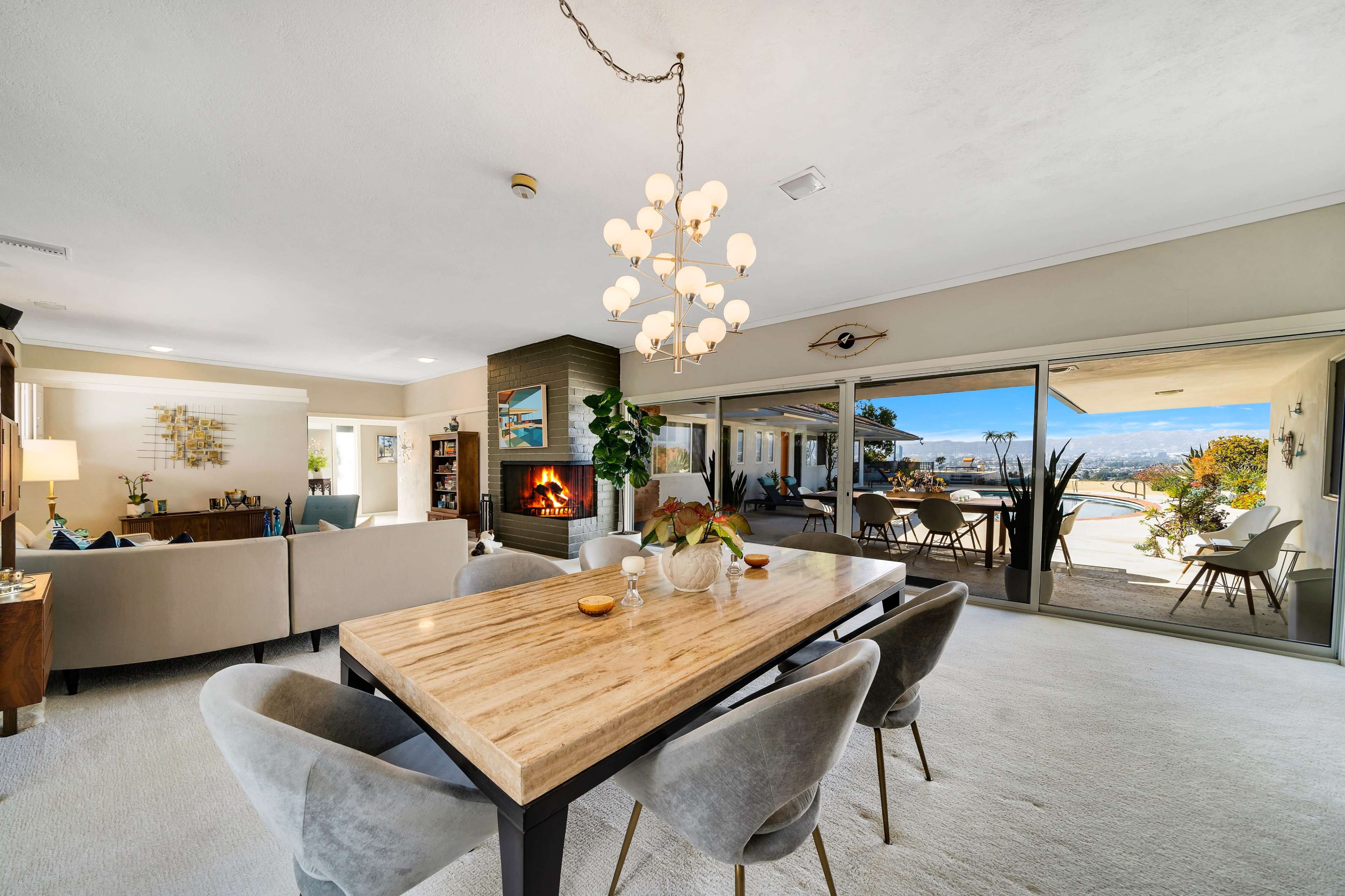 The image shows a modern living and dining area with a wooden table surrounded by chairs, a cozy fireplace, and large glass doors open to a patio with a view.