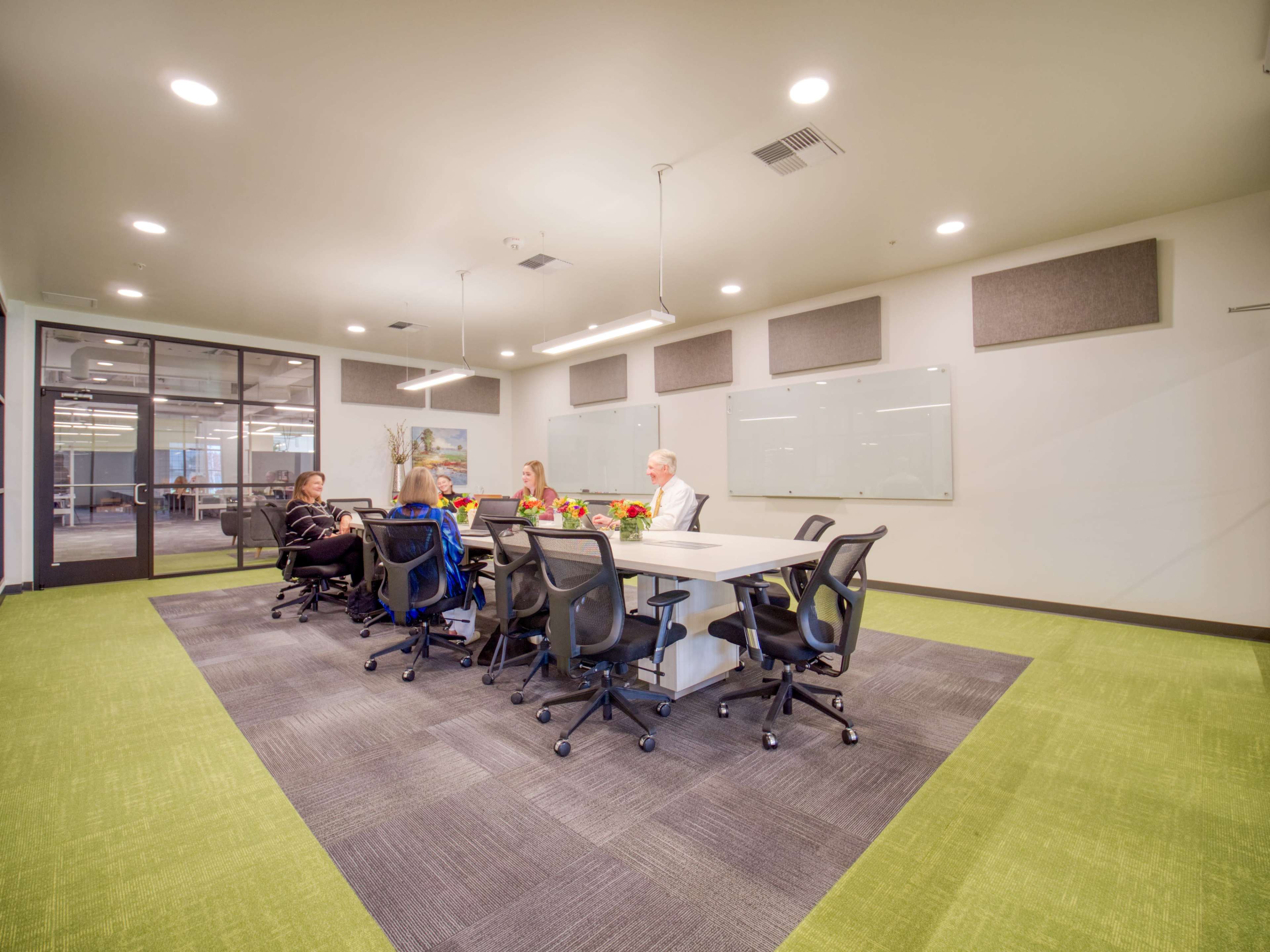 A group of five people sits around a rectangular conference table in an office meeting room with green carpet and white walls.